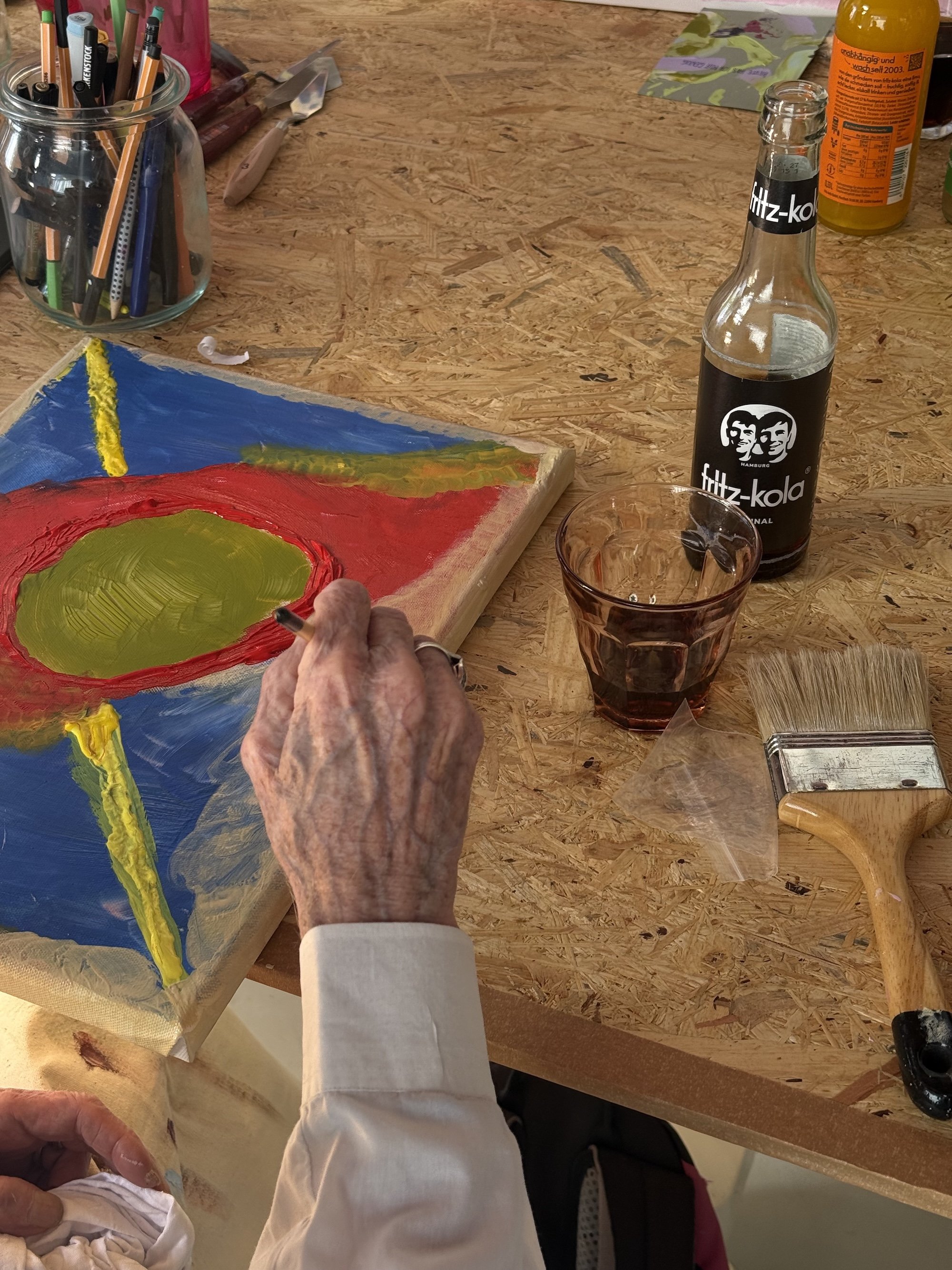 Der Tisch ist mit Malutensilien wie Pinsel, Farben und einem Glas mit roter Flüssigkeit bedeckt. Im Hintergrund sind Flaschen und andere Materialien sichtbar. Eine Person malt auf einer Leinwand mit bunten Farben.