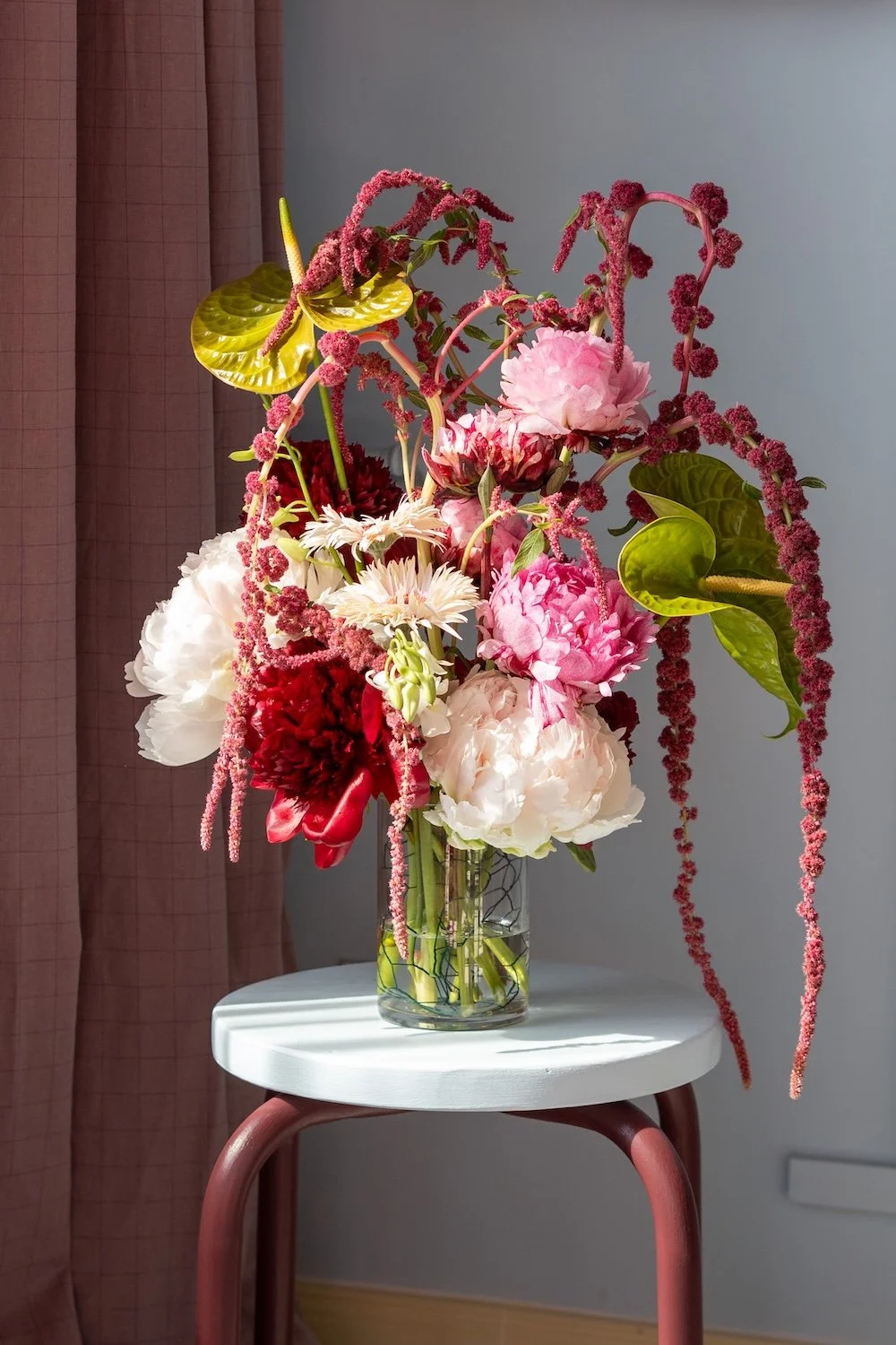 Vasen mit bunten Blumen, darunter Pfingstrosen, Gerbera und Astilben, auf einem weißen Tisch mit rotem Stuhl im Hintergrund.