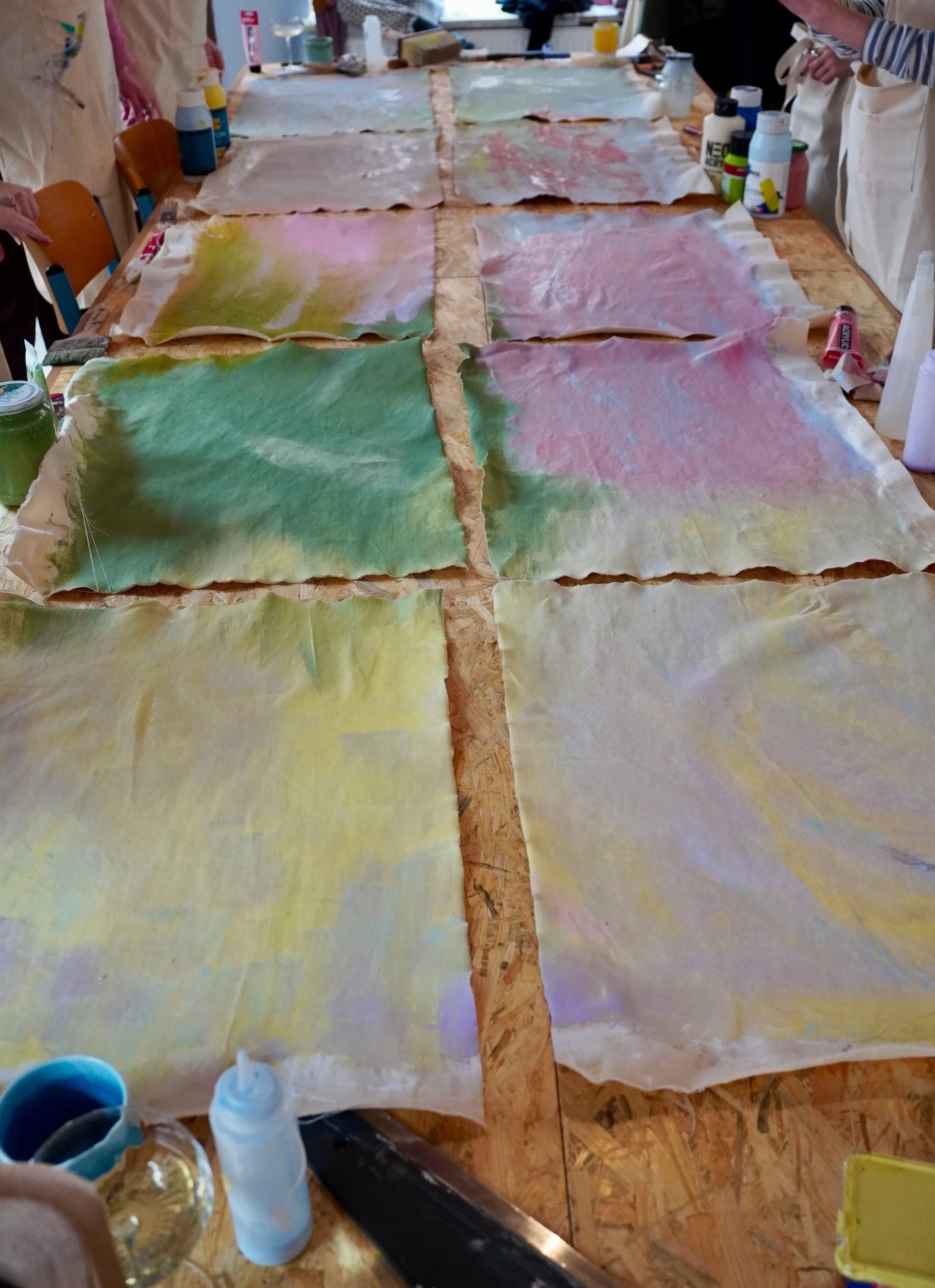 Mehrere bunte, halb getrocknete Papierstücke liegen auf einem Holztisch, in der Umgebung befinden sich Bastelmaterialien und Personen beim Basteln.