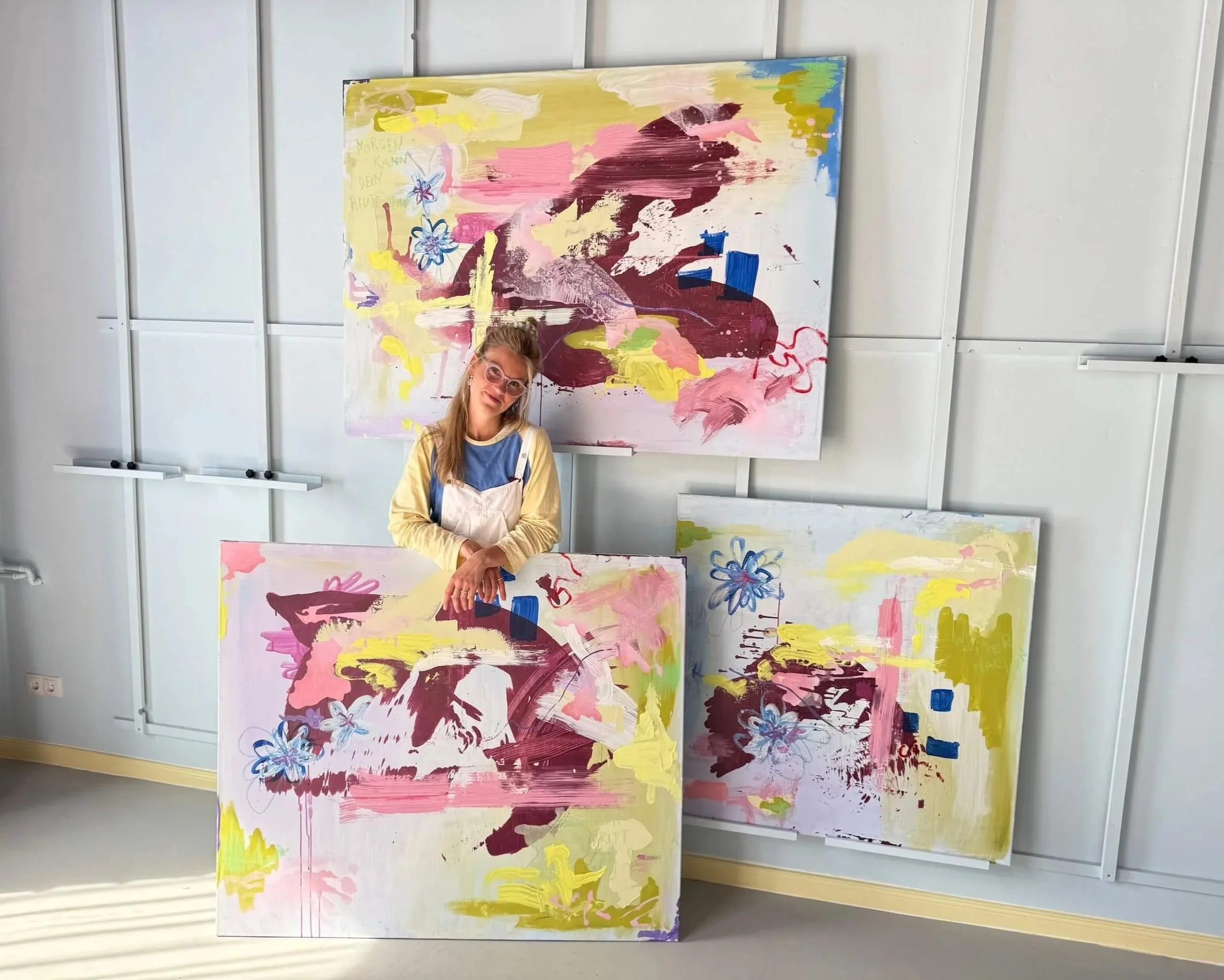 Ein junges Mädchen mit Brille und blonden Haaren steht vor farbenfrohen abstrakten Gemälden, die an einer Wand im Kunststudio hängen.