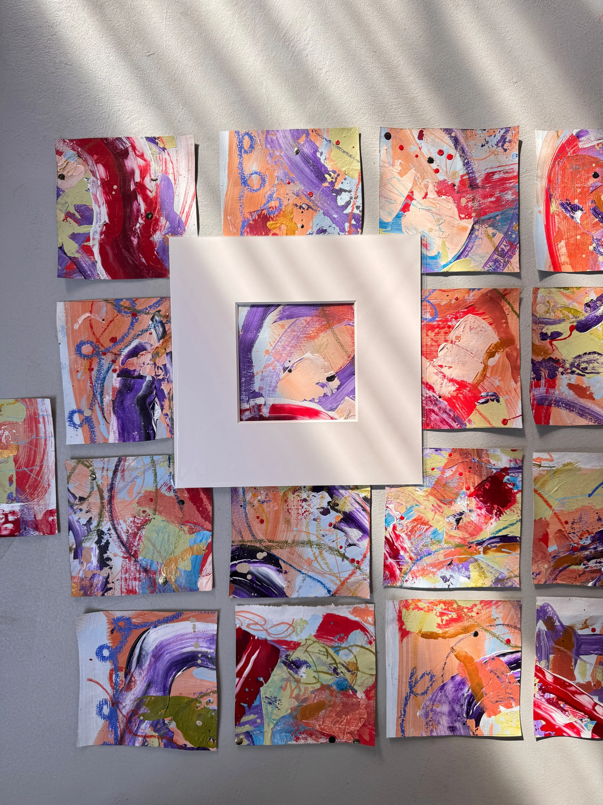 Mehrere kleine bunte Gemälde in abstraktem expressionistischen Stil, an einer weißen Wand angebracht, mit lebhaften Farben wie Rot, Lila, Orange, Gelb und Blau, einige Gemälde sind in weißen Rahmen eingefasst.