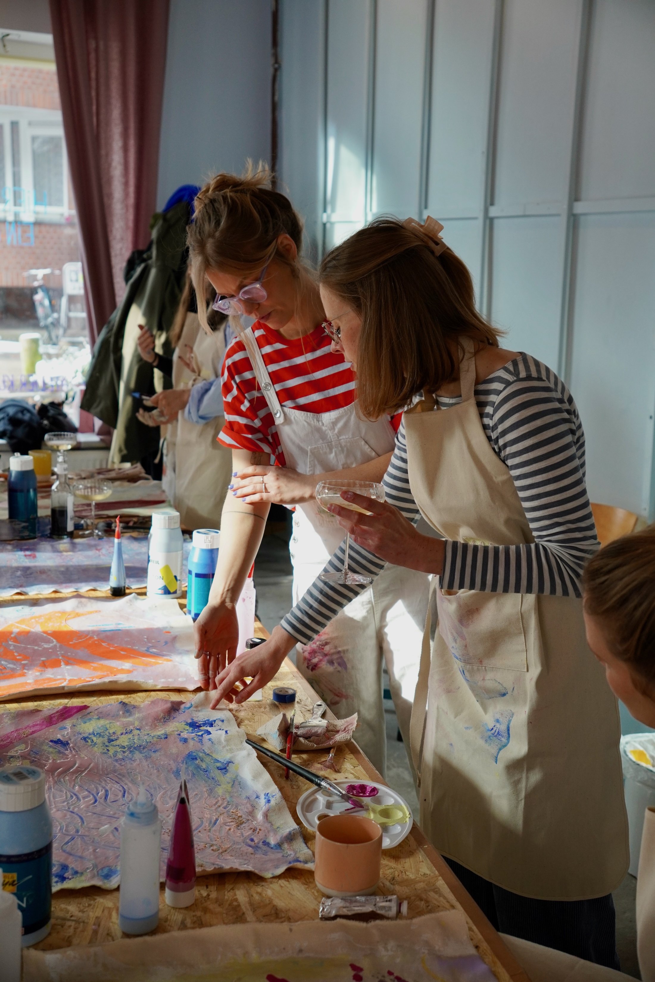 Zwei Frauen beim gemeinsamen Malen und Sehen eines abstrakten Kunstwerks auf einer großen Leinwand, umgeben von Farben und Pinsel in einem Atelier.