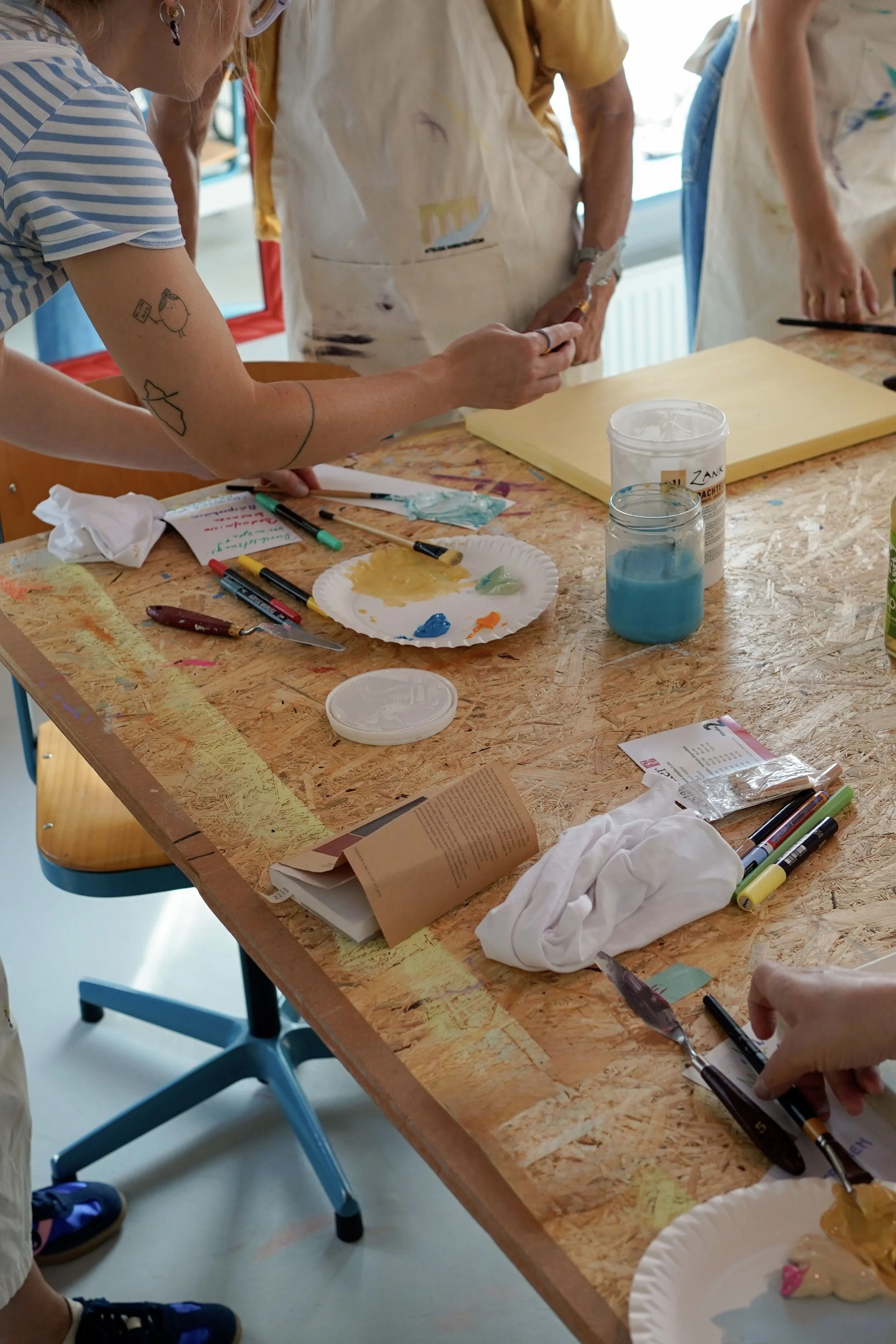 Menschen beim Basteln oder Malen in einem Raum, mit Bastelmaterialien auf einem Holzarbeitstisch, darunter Farben, Marker und Papier.
