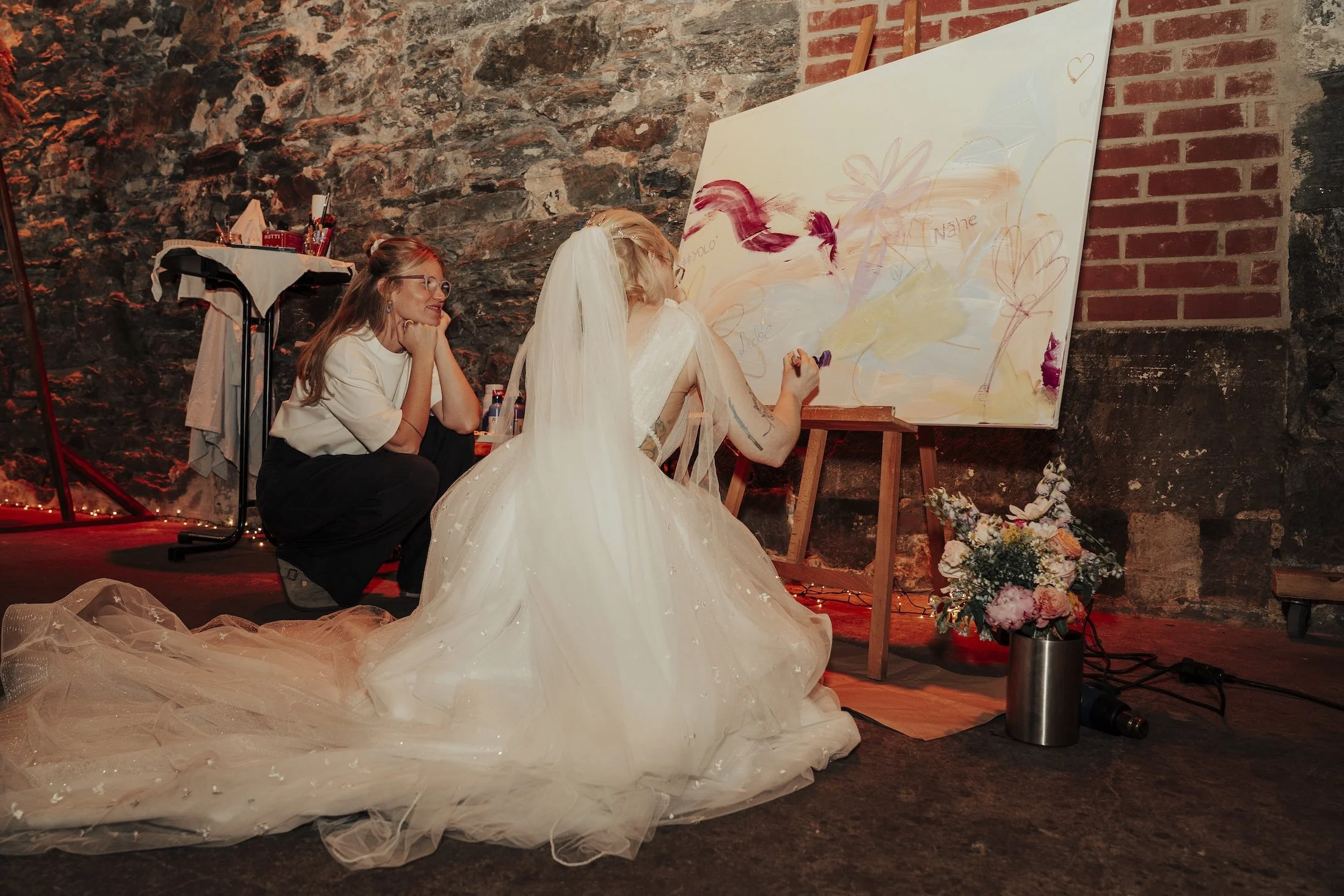 Eine Braut in weißem Kleid und Schleier beim Malen auf einer großen Leinwand, neben einer Frau, die zuschaut. Die Szene scheint eine kreative Hochzeit oder Feier zu sein, mit Blumen und Dekoration im Hintergrund.