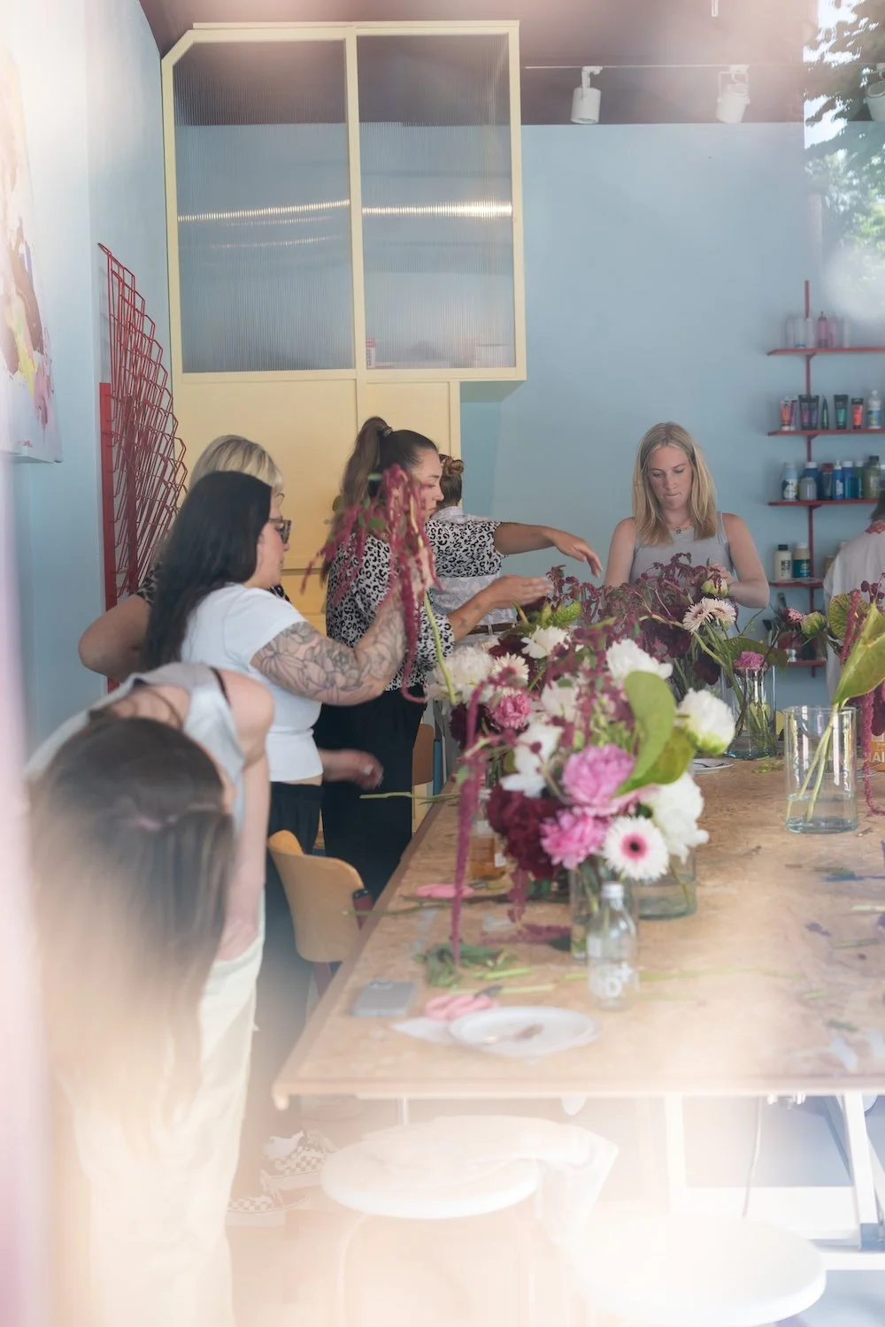Mehrere Frauen gestalten Blumenarrangements auf einem Tisch mit bunten Blumen in Vasen, in einem hell erleuchteten Raum.