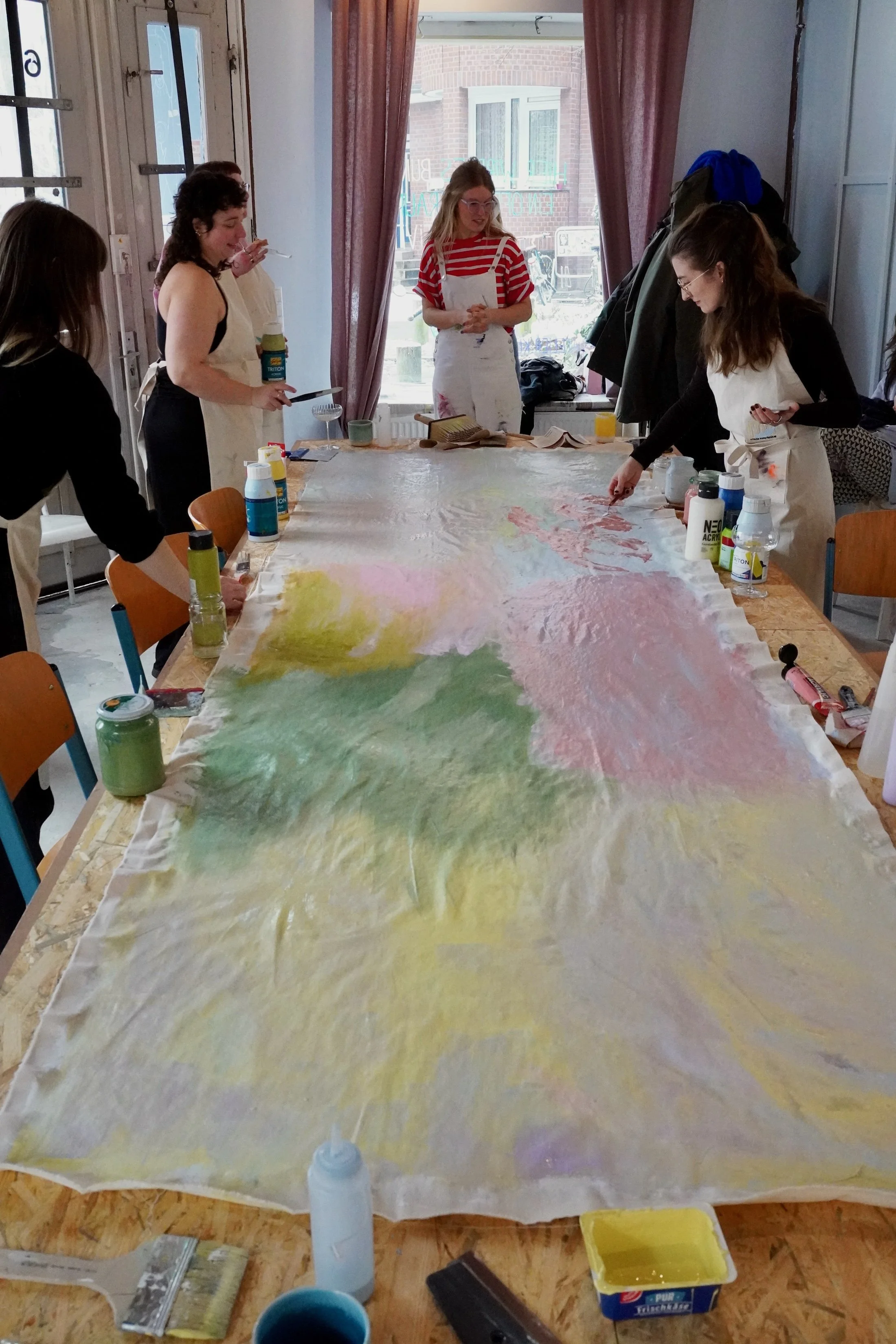 Mehrere Menschen, Frauen, die an einem großen papierähnlichen Kunstwerk arbeiten, das auf einem Tisch aus Holz liegt. Sie verwenden Farben und Pinsel, um das Kunstwerk zu gestalten. Im Hintergrund befindet sich eine Fensterscheibe mit Vorhängen, durc