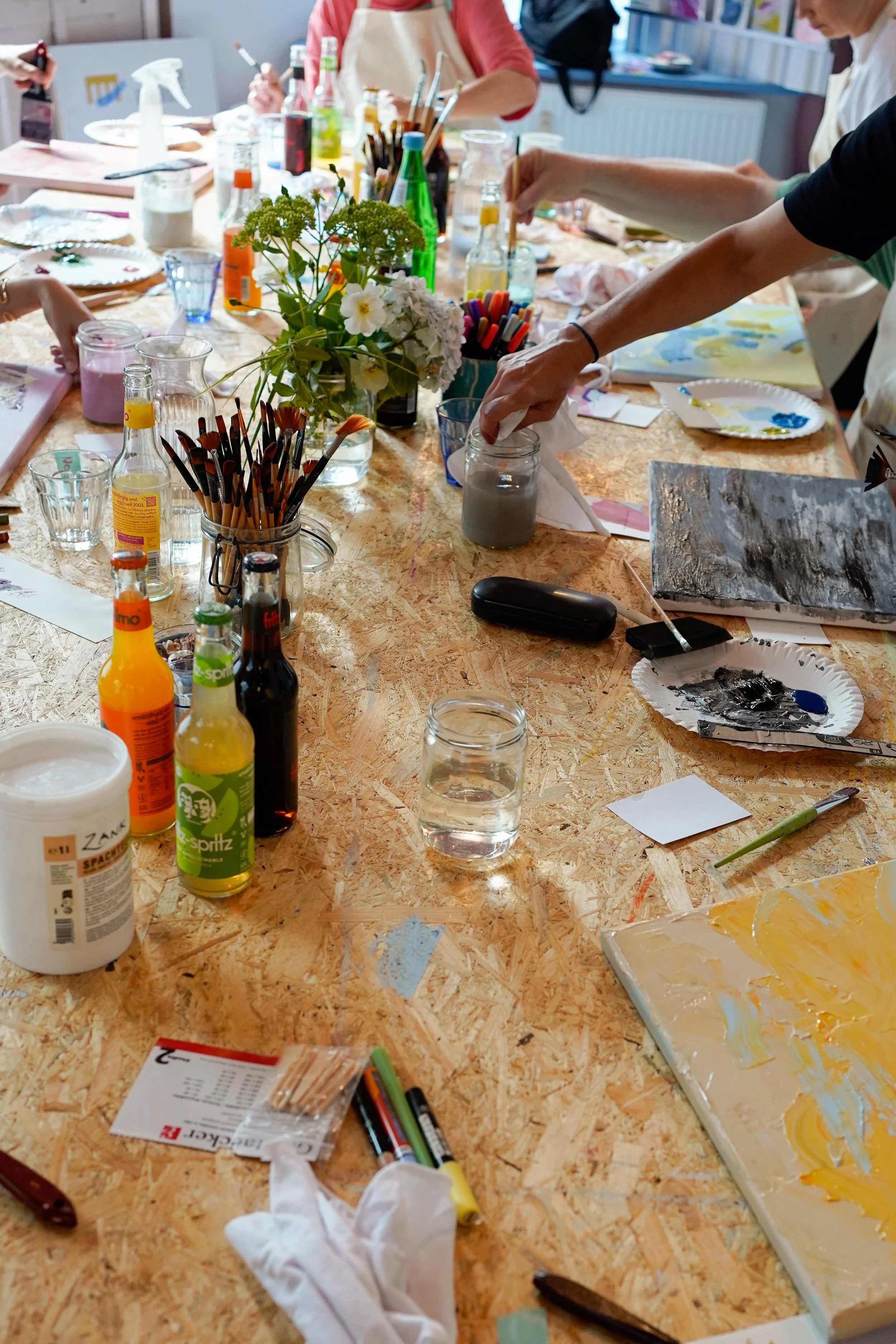 Ein Tisch, auf dem Malutensilien wie Pinsel, Farben, eine Wasserflasche, Pappteller mit Farbe, eine Blume in einer Vase und mehrere Getränkeflaschen und Gläser stehen. Es sind Personen beim Malen und Basteln aktiv.