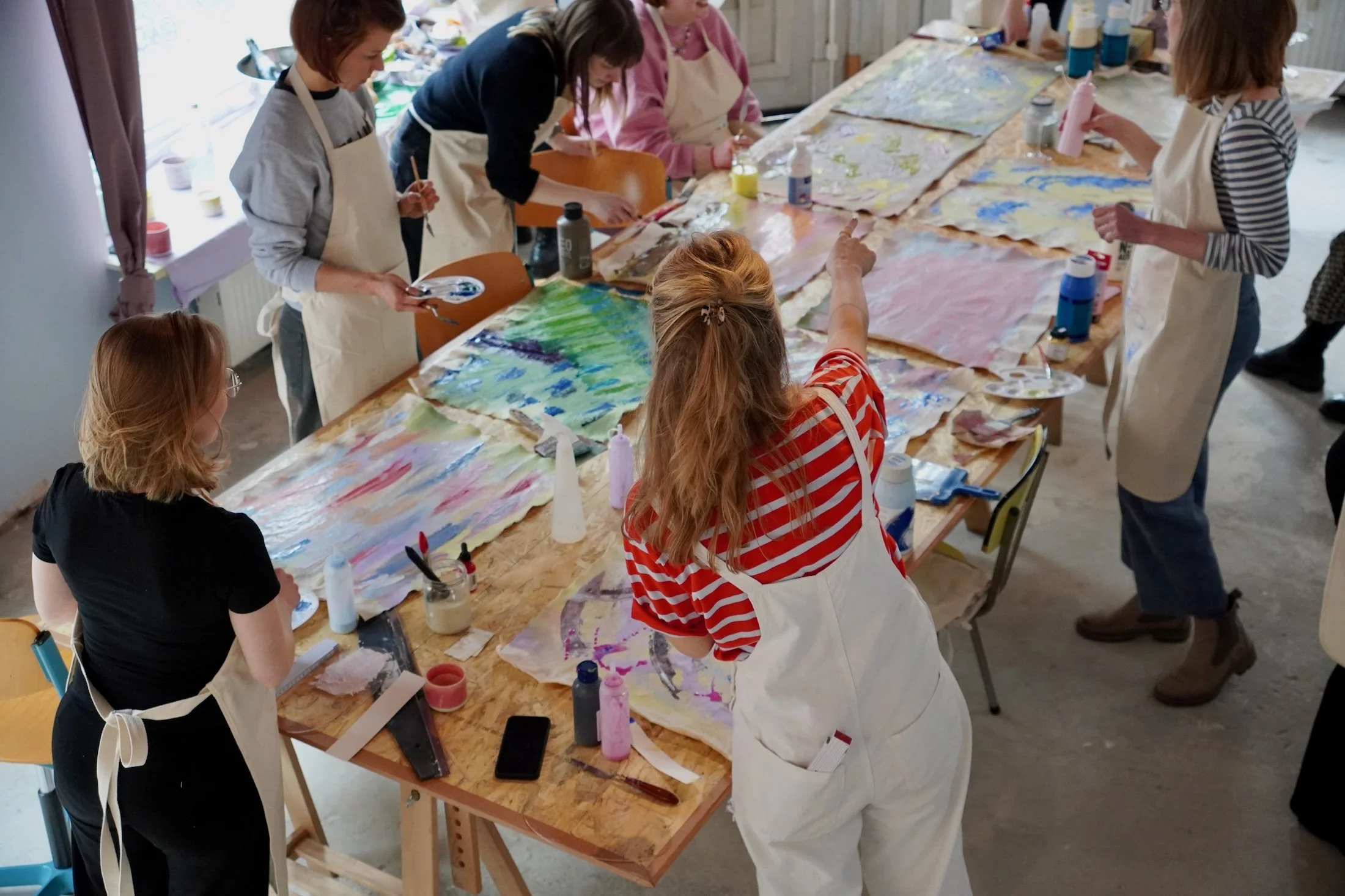 Menschen beim gemeinsamen Malen oder Basteln in einem kunstvollen Workshop. Verschiedene Kunstwerke auf dem Tisch, Farben und Materialien liegen verteilt. Die Beteiligten tragen Schürzen, um ihre Kleidung zu schützen.