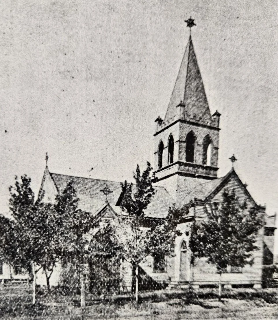 Black and white photo of a church with a tall steeple topped with a star, trees surrounding the church yard, and a fence in the foreground.