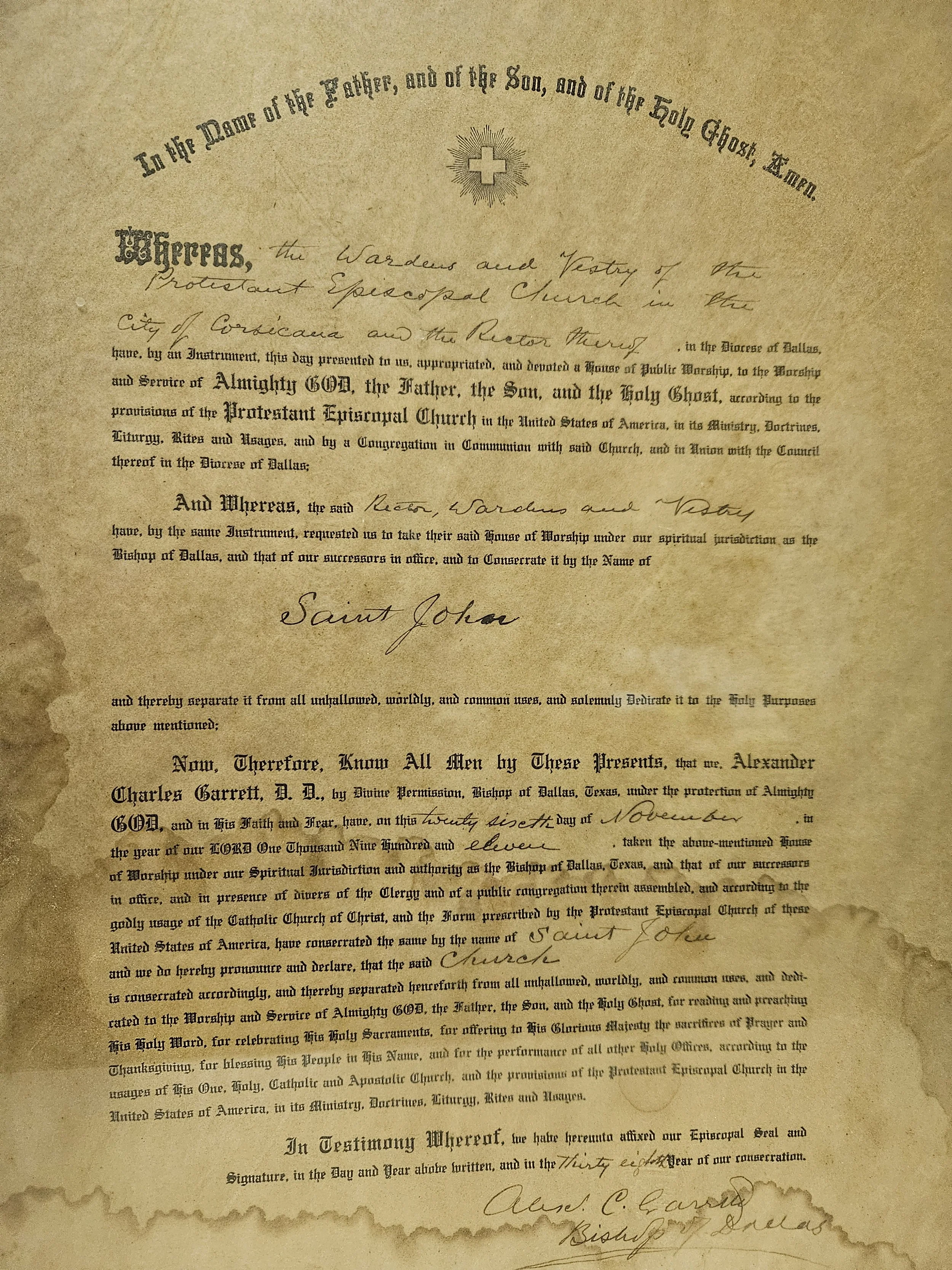 Historical document with ornate text, signatures, and a decorative cross at the top center. Paper appears aged with burnt edges.