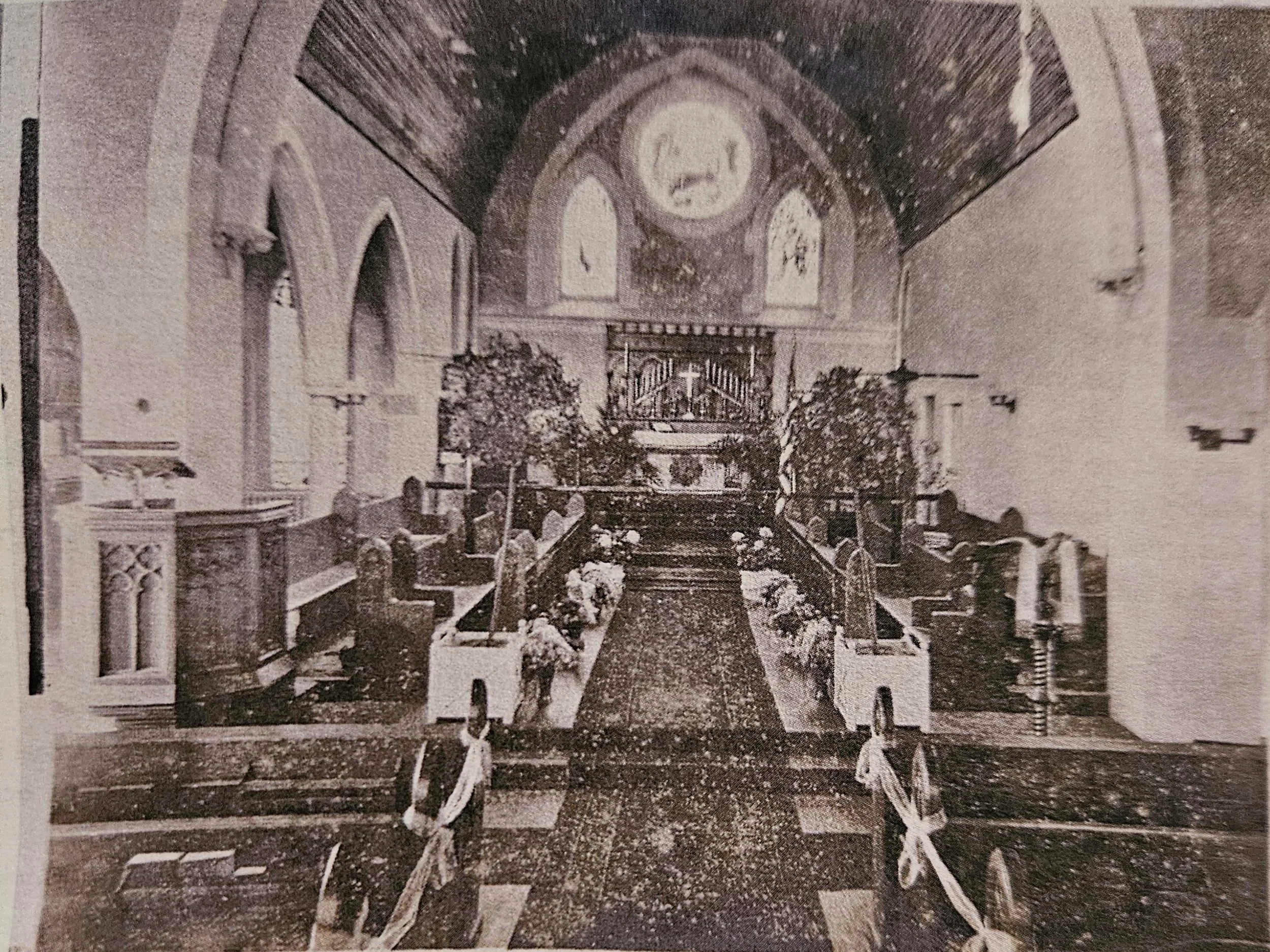 A vintage photograph of the interior of a church with wooden pews, flowers, and a simple altar at the front, featuring stained glass windows and religious symbols.