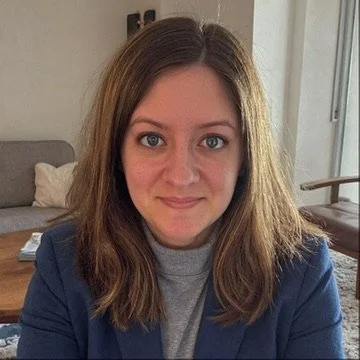 A young woman with shoulder-length brown hair, wearing a blue blazer and gray top, sitting indoors with a sofa and window in the background.