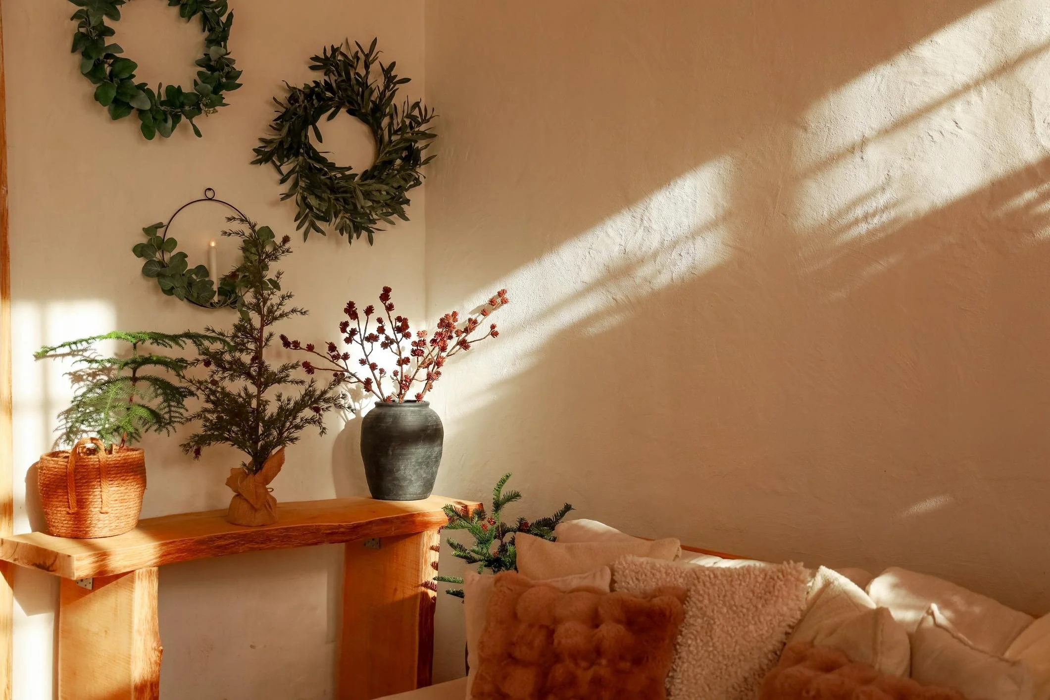 A cozy corner of a living room with a wooden table, potted plants, and wreaths hanging on the wall. Sunlight creates shadows on the beige textured wall.