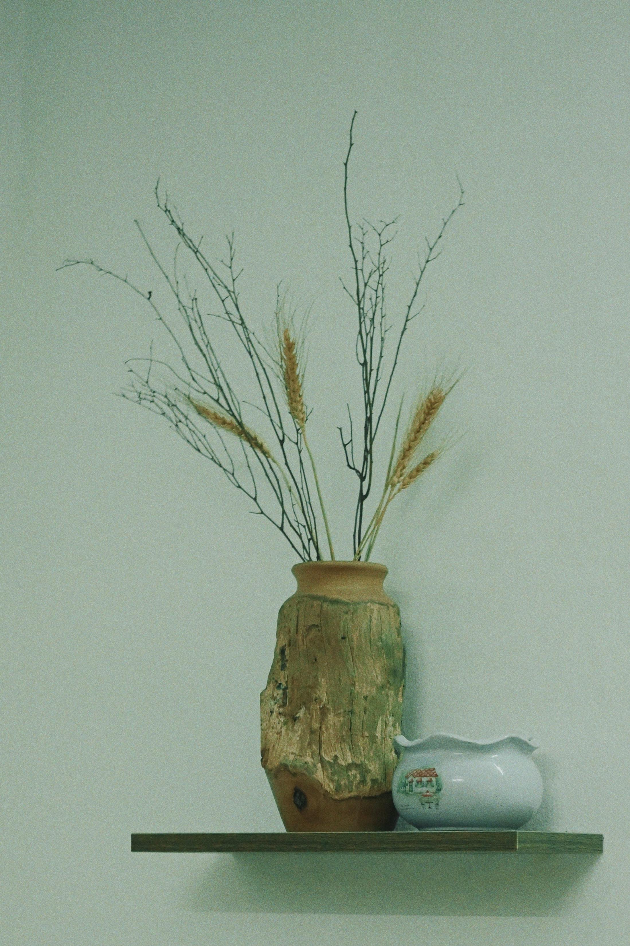 A ceramic vase with dried grass and twigs, placed on a wooden shelf above a ceramic container with painted houses.