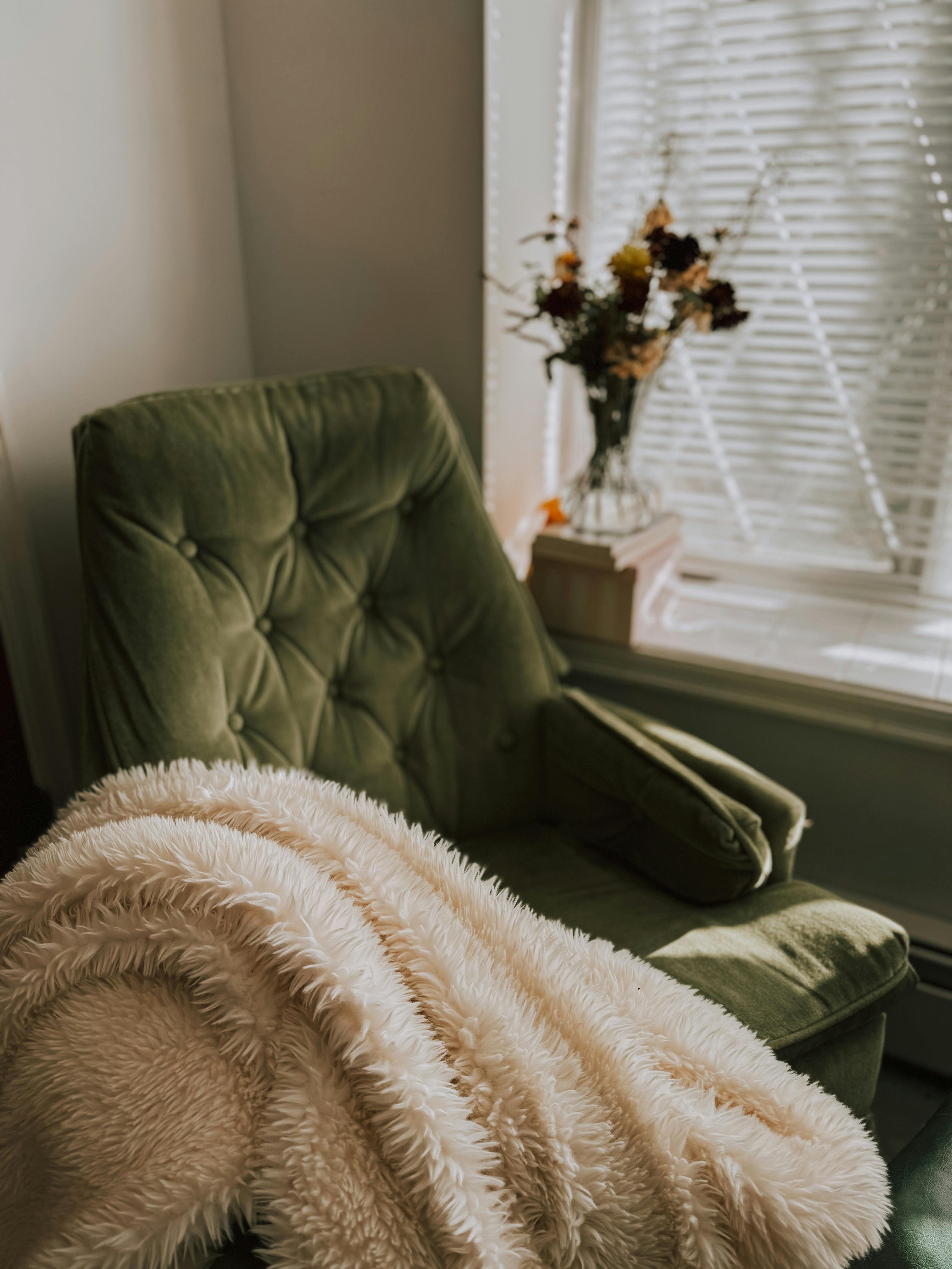 Close-up of a cozy corner with a plush off-white faux fur blanket, part of a green velvet armchair, a window with white blinds, and a small table with a vase of dried flowers.