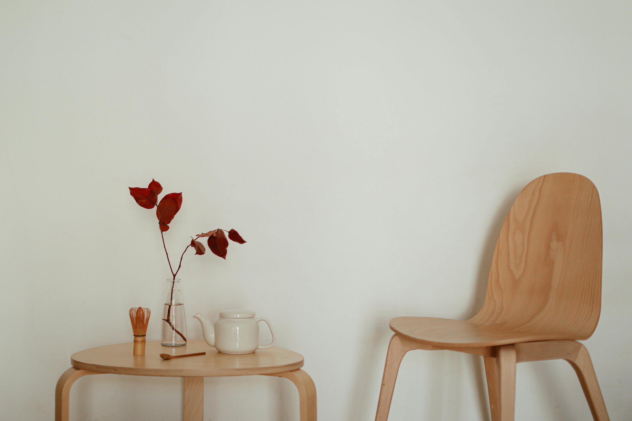 A light-colored wooden chair with a curved backrest and rounded seat is placed next to a small round wooden table. On the table, there is a glass vase with red leaves, a small wooden object, a white teapot, and a cup. The background is a plain, light-colored wall.