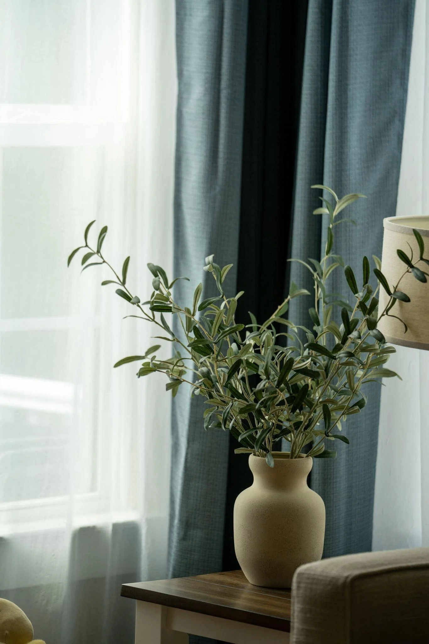 A beige decorative vase with a round body and slender neck, placed on a wooden table, contains green leafy branches. In the background, there are sheer white curtains and darker gray curtains, with soft natural light coming through a window.