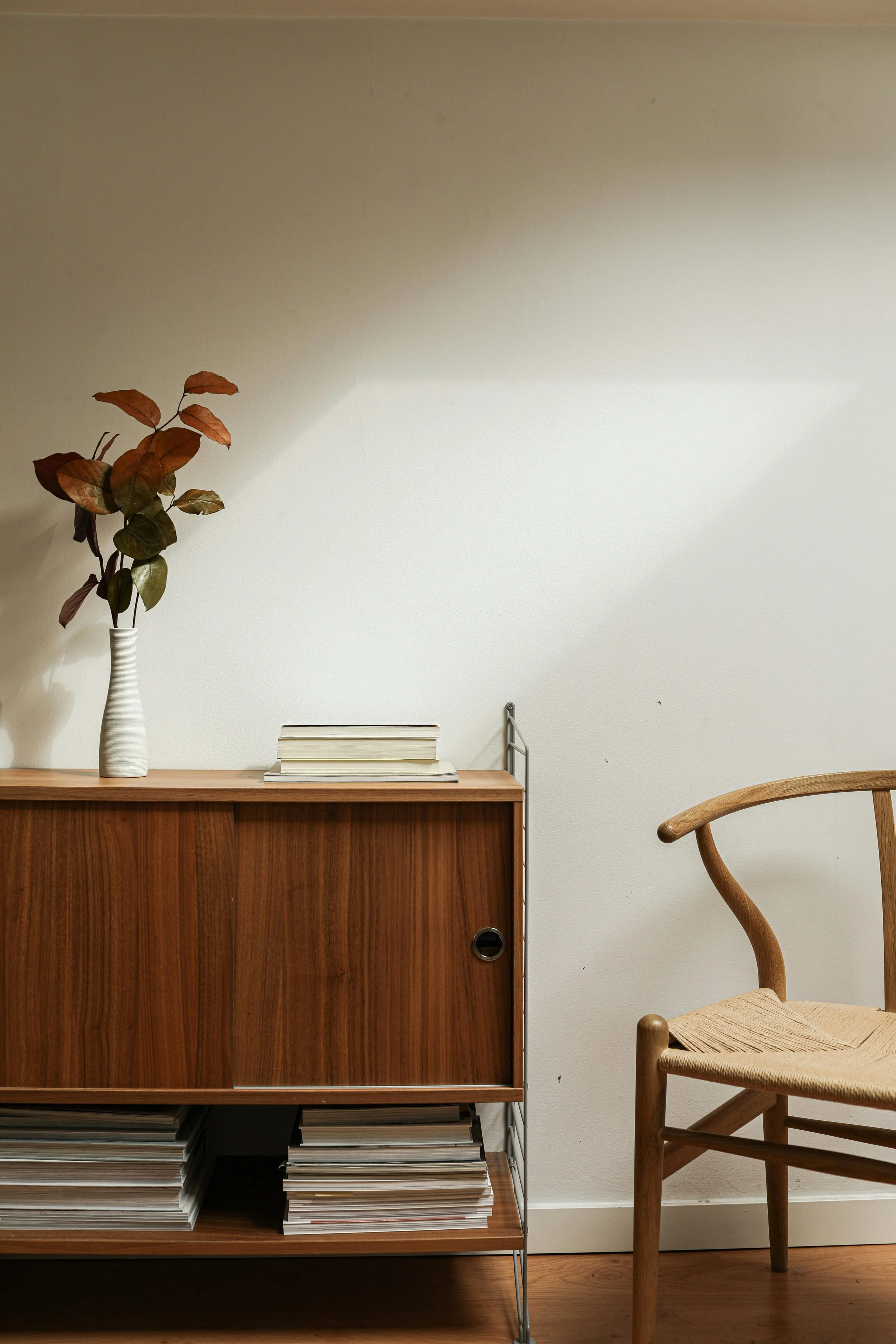 A wooden cabinet with a stack of magazines or books on top, a white vase with a branch of reddish-brown leaves, a white wall, and a wooden chair with a woven seat.