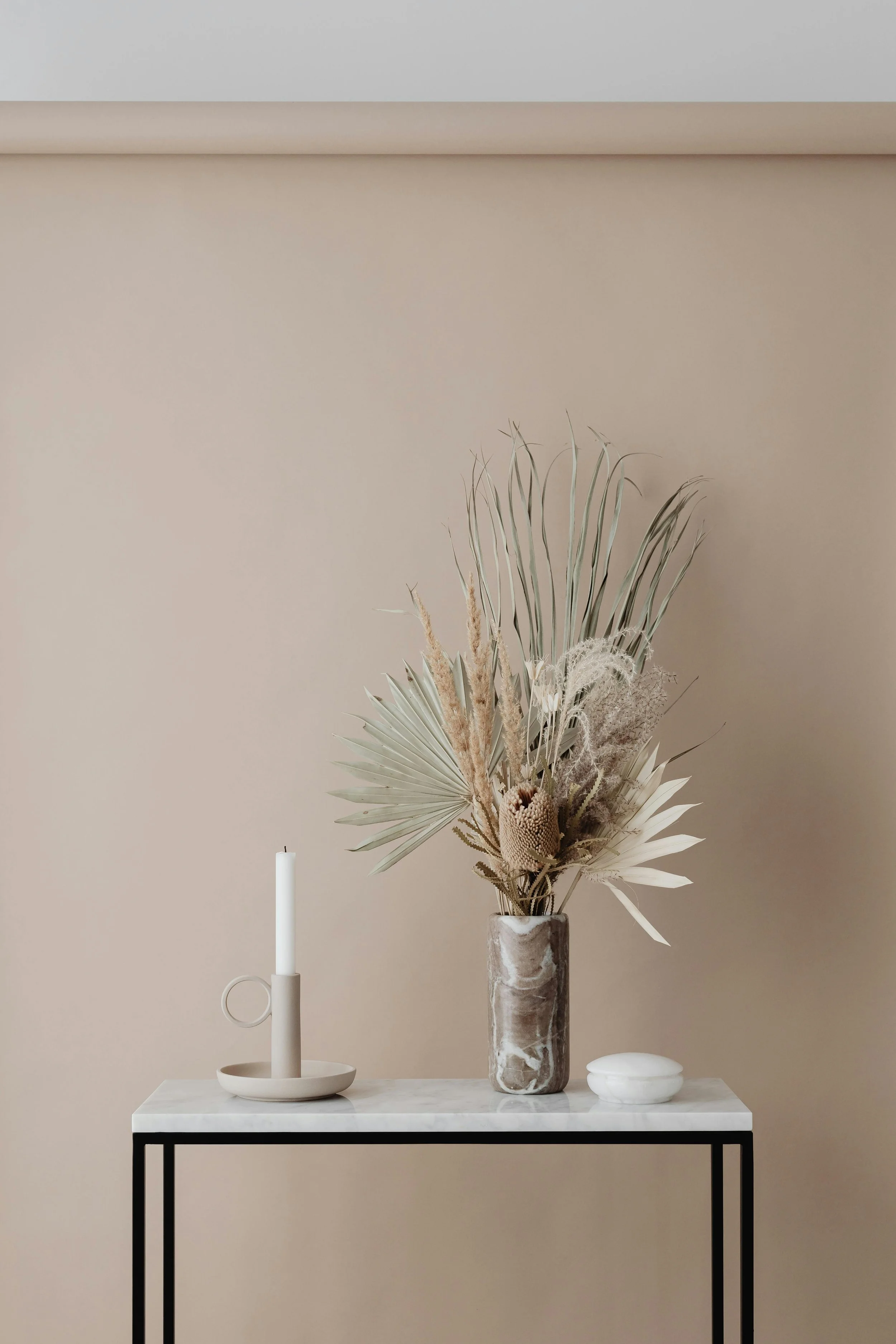 A minimalist floral arrangement in a marble vase, placed on a white table with black legs. The arrangement includes dried palm leaves and other dried flowers. To the left of the vase, there is a white candlestick with a tall, white, unlit candle. To the right of the vase, there is a small white decorative object. The background wall is plain and beige.