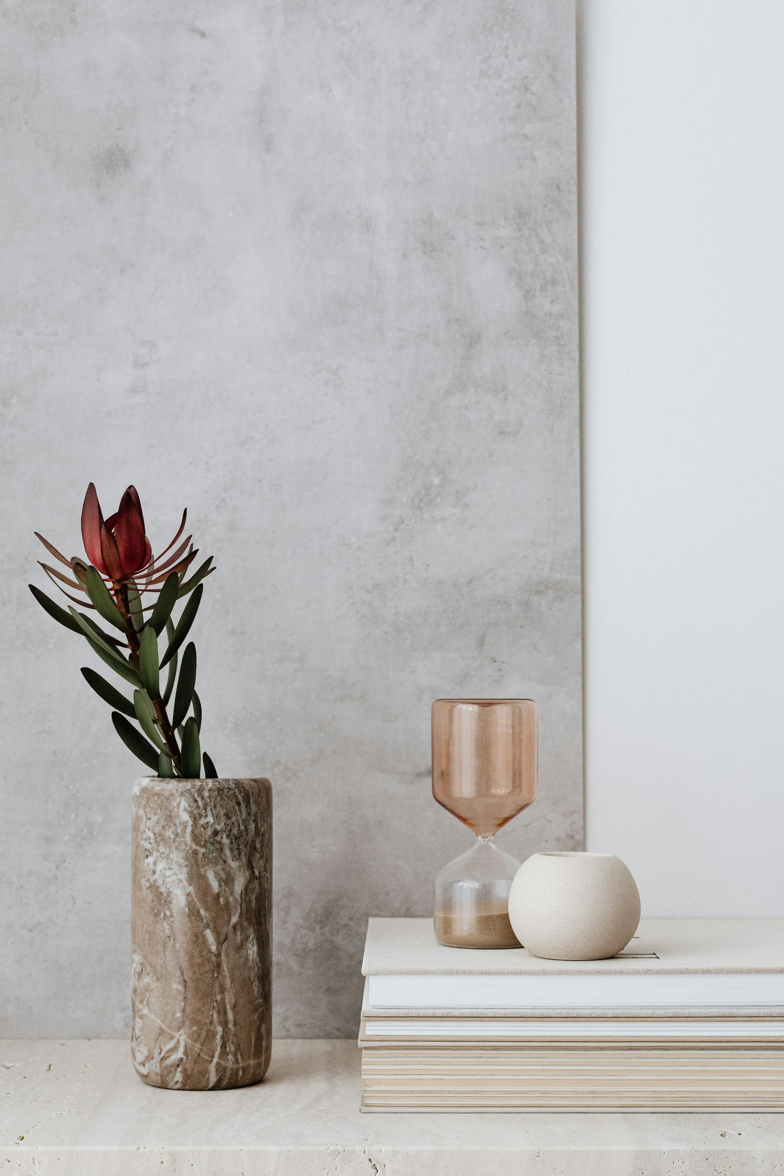 Decorative arrangement featuring a tall marble-based vase with a green plant and red flower, a pinkish glass candle holder, a geometric white vase, and a stack of white books with a minimalist design on a white surface against a textured gray wall.