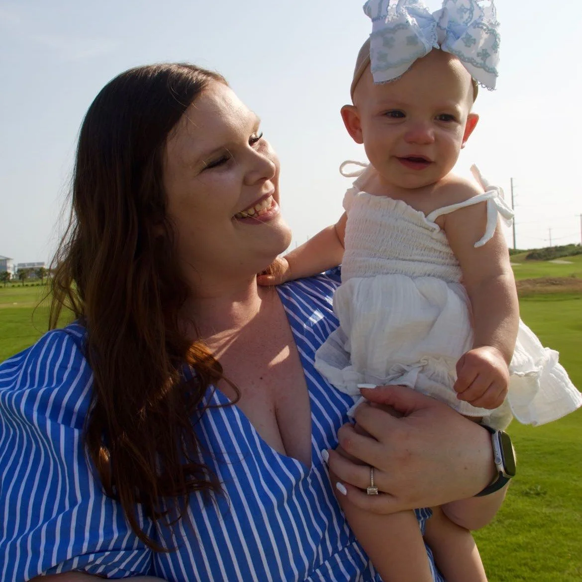 A woman holding a young girl outdoors on a sunny day, both smiling and enjoying the moment.