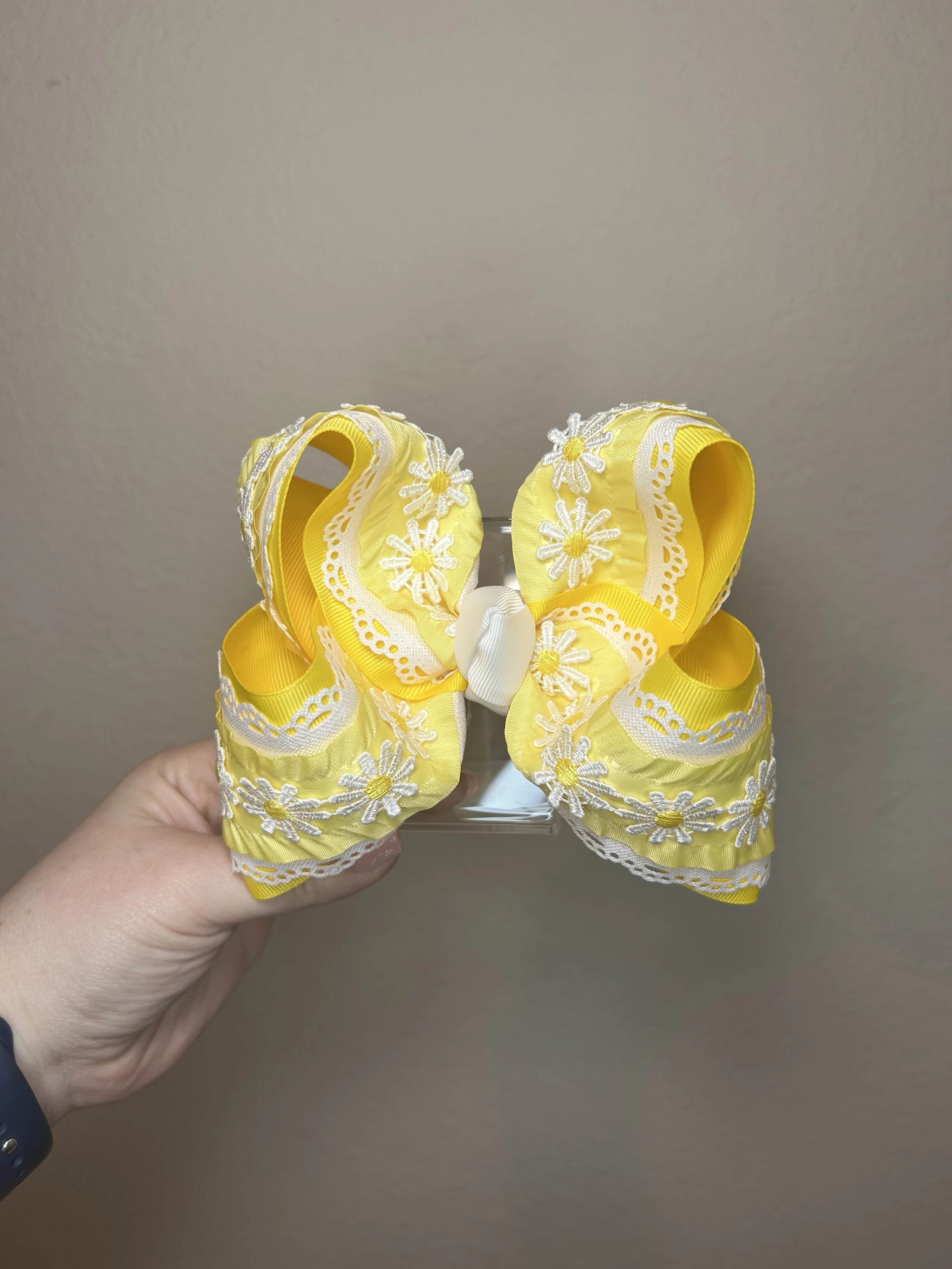 Yellow fabric bow with white embroidered daisies and lace trim, held by a person's hand against a plain beige wall.