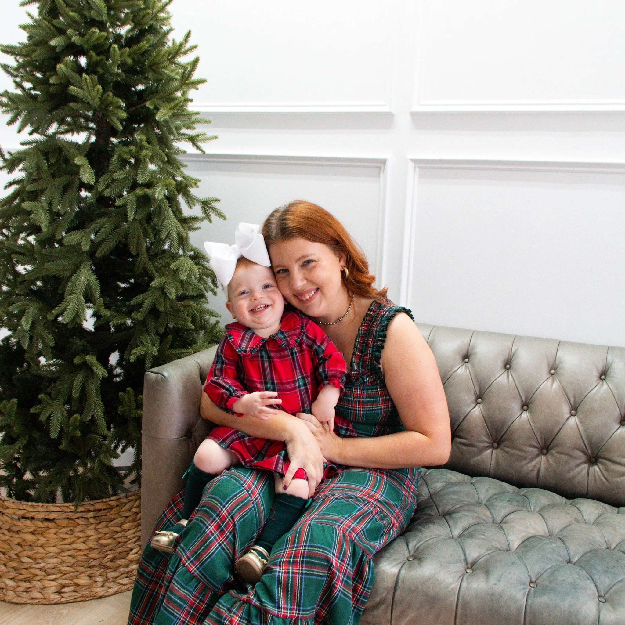 A woman and a young girl sitting on a gray leather sofa next to a Christmas tree, both smiling and dressed in matching red and green plaid outfits.