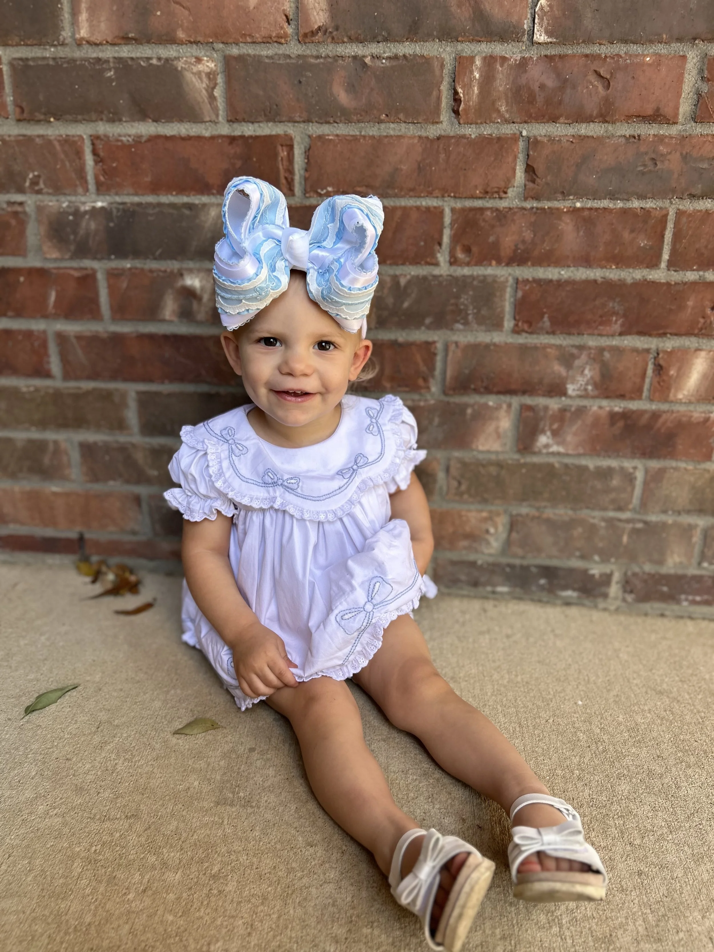 A young girl sitting on the ground in front of a brick wall, wearing a white dress with ruffled sleeves and embroidery, white sandals, and a large white and blue bow headband, smiling at the camera.