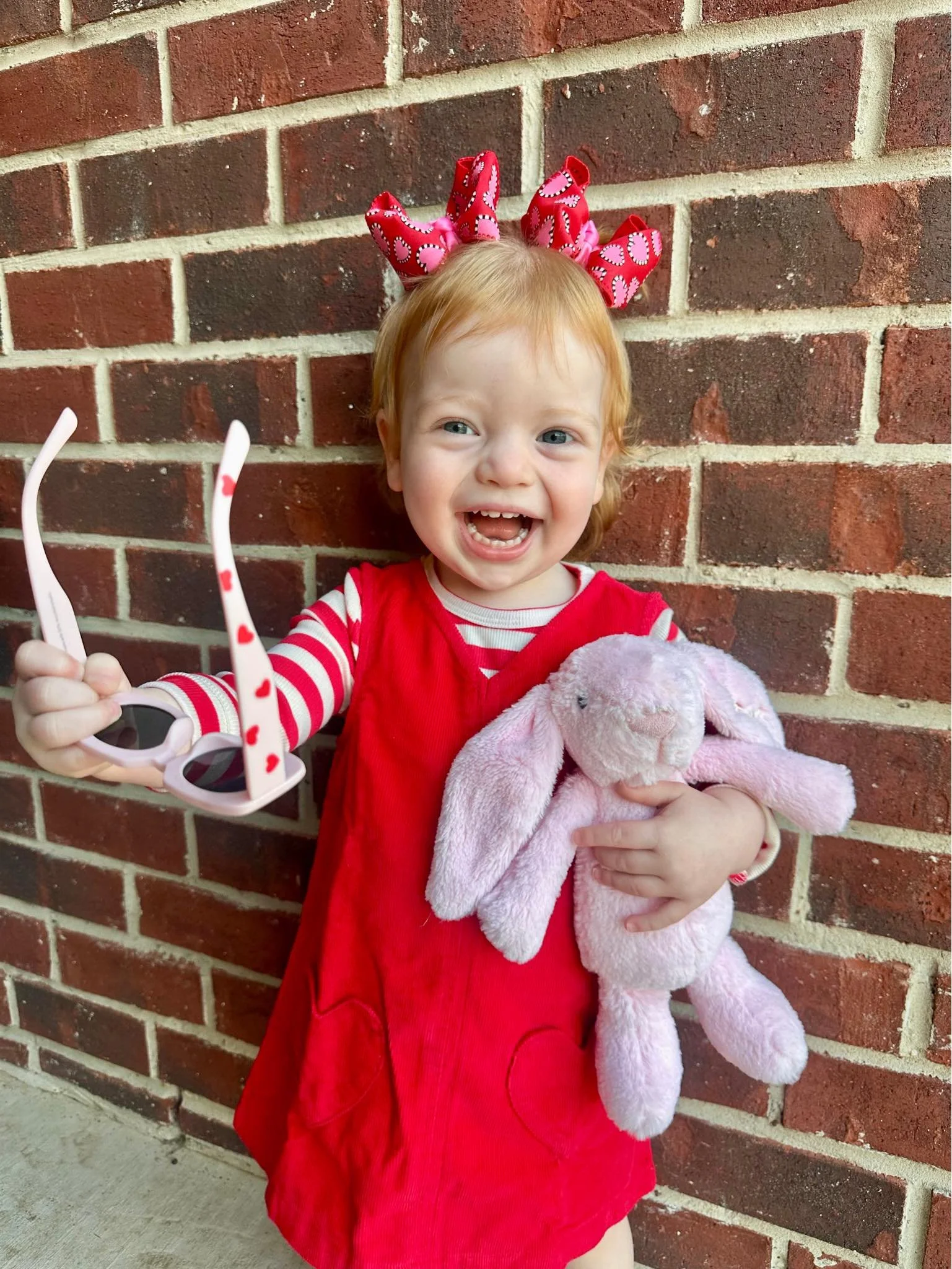 A young girl with red hair, wearing a red dress with white and red striped long sleeves, holding pink sunglasses with heart patterns in one hand and a pink plush bunny in the other, standing against a brick wall.