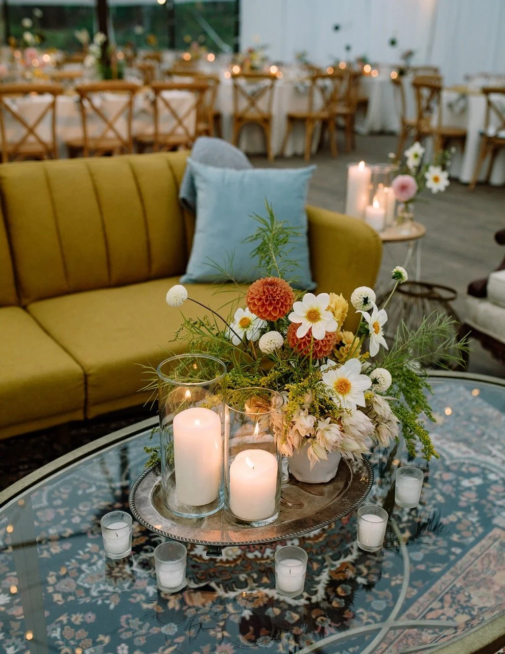 A cozy indoor event space with a yellow sofa, a blue pillow, and a glass coffee table. The table holds a floral centerpiece with white, orange, and yellow flowers in a white vase, surrounded by candles in glass holders, creating a warm ambiance. In the background, there are round tables with wooden chairs, set for a gathering.