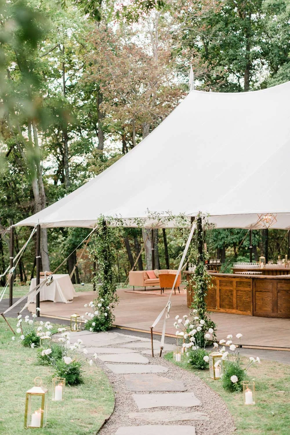 Outdoor wedding or event setup with a large white tent, decorated with greenery and white flowers, surrounded by lanterns and a stone pathway.
