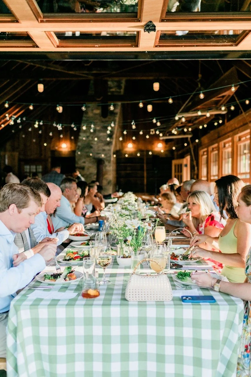 People gathered for a large outdoor meal under a rustic wooden barn with string lights, with a long table decorated with flowers and drinks.
