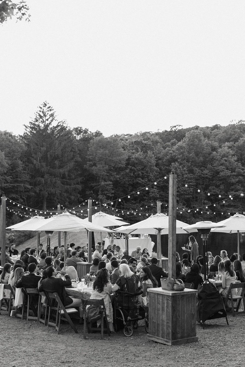 An outdoor gathering with people sitting at tables under string lights and umbrellas, with a wooded hillside in the background.