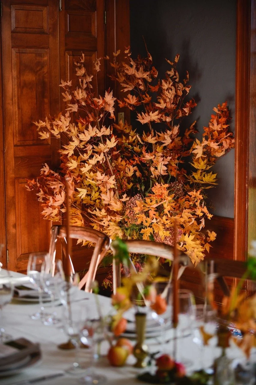 A table decorated with glassware, plates, and apples, with a large arrangement of autumn leaves and flowers in the background.