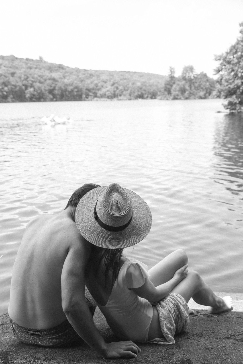 A couple sitting by the lakeshore, with the woman leaning into the man, both wearing swimwear, and the woman wearing a wide-brimmed hat. In the background, a person can be seen kayaking on the lake, surrounded by wooded hills.