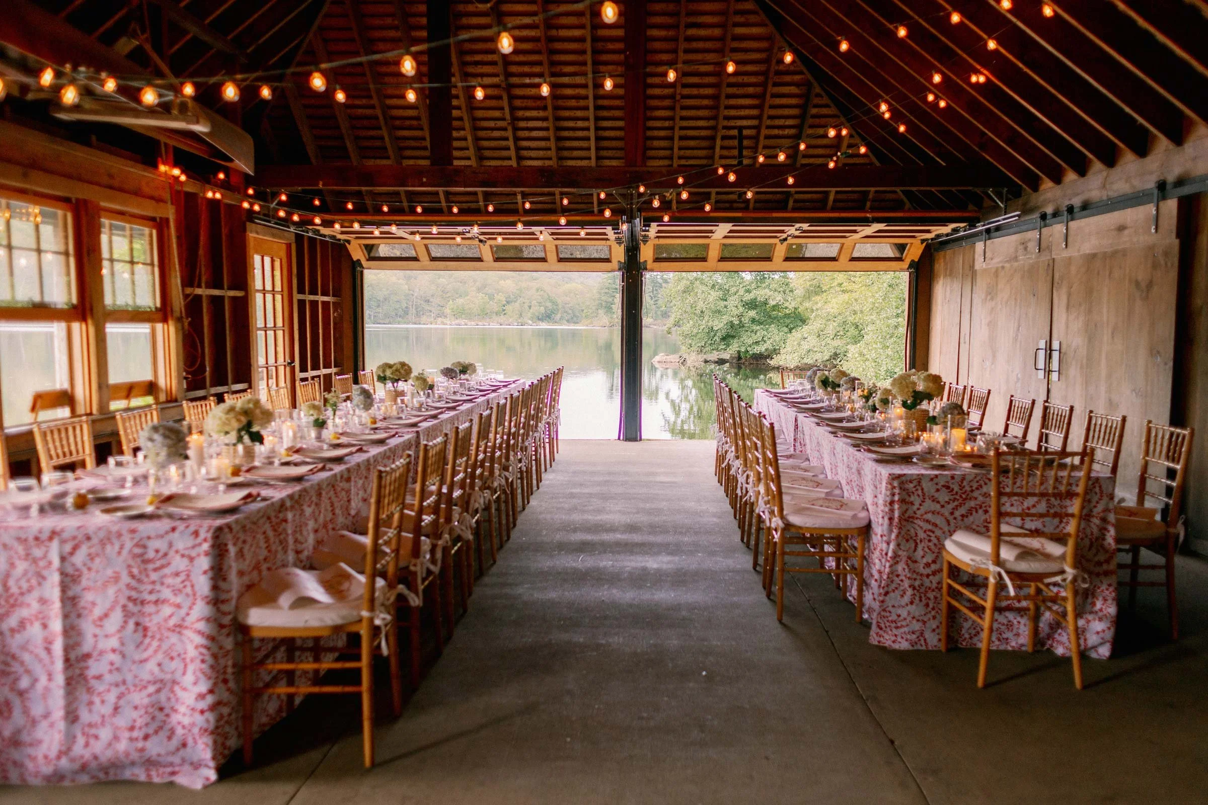 Long dining tables with floral tablecloths and floral centerpieces inside a rustic barn with string lights and an open view of a lake and trees outside.