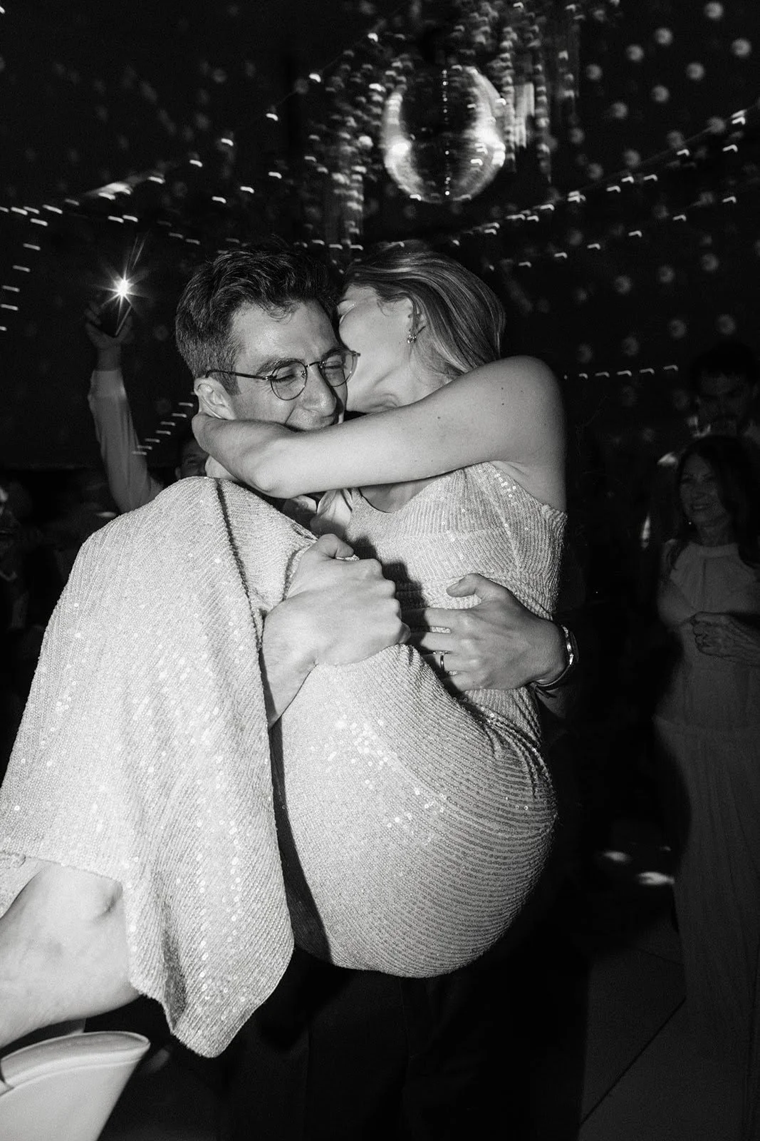 A black and white photo of a couple dancing closely at a party, with a disco ball overhead and other guests in the background.