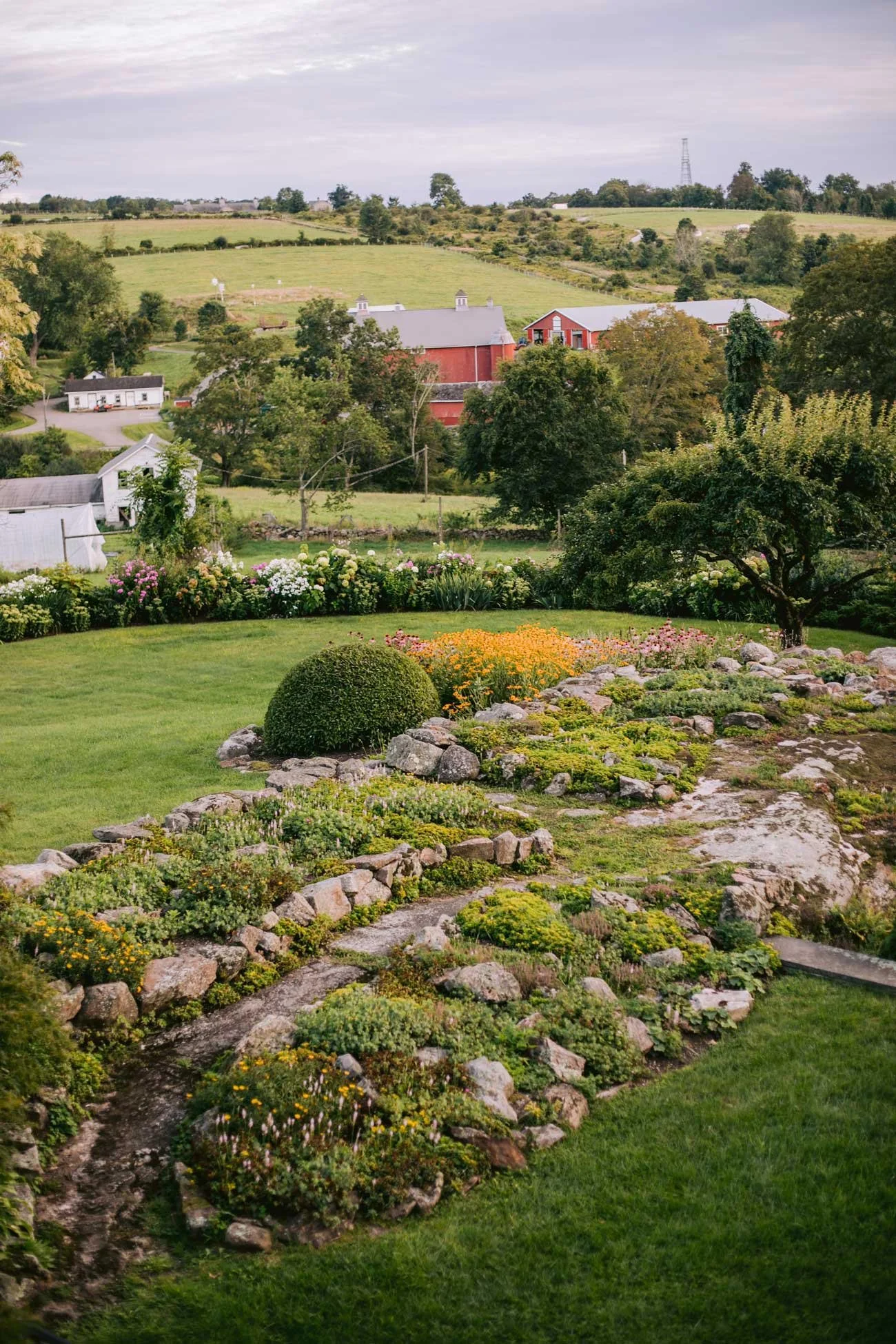 A rural landscape featuring a garden with flowering plants, trees, a small path made of rocks, rolling hills with green fields, and farm buildings in the distance under a cloudy sky.