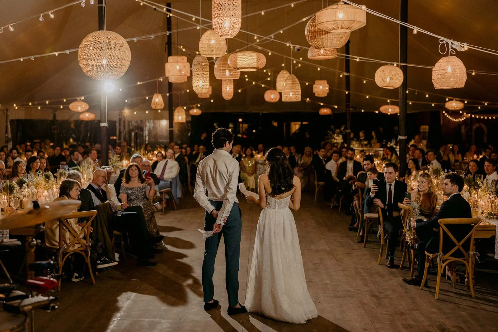 A couple is standing in front of seated guests during a wedding reception under hanging wicker lanterns and string lights in a large tent.