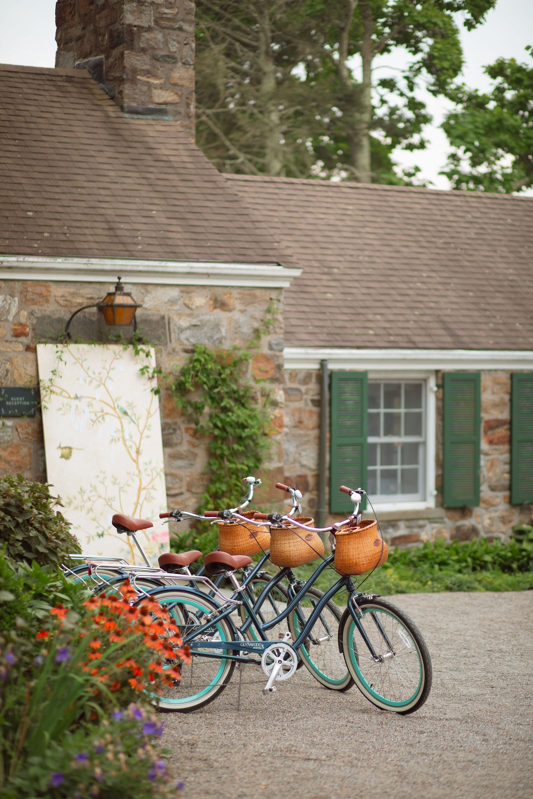 Four bicycles with baskets parked in front of a stone house with a window with green shutters and a flowered garden.