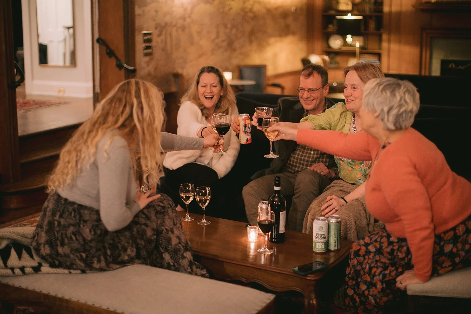 Group of six friends having drinks and toasting in a cozy, warmly lit living room.