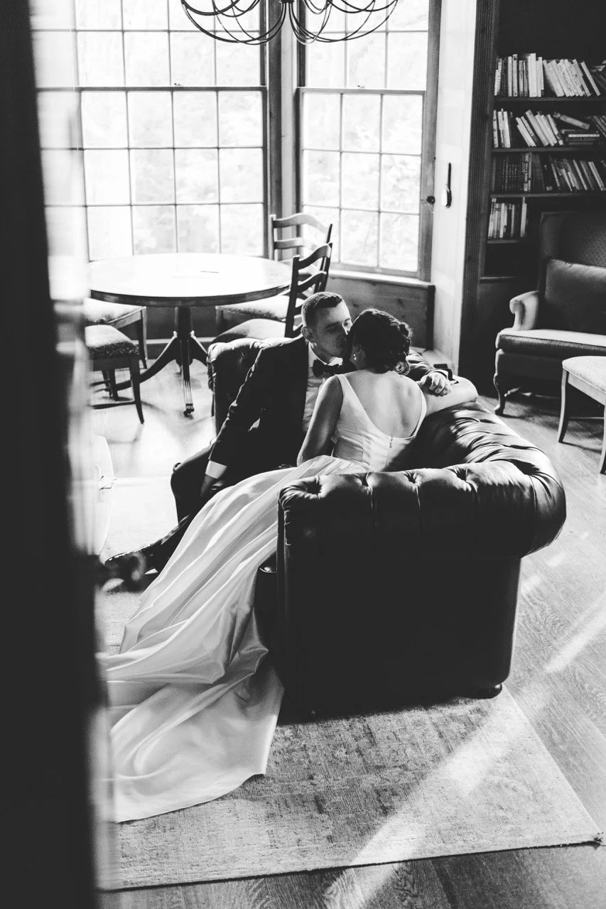 A black-and-white photo of a bride and groom sharing a moment on a sofa in a cozy room with large window, bookshelf, and fireplace.