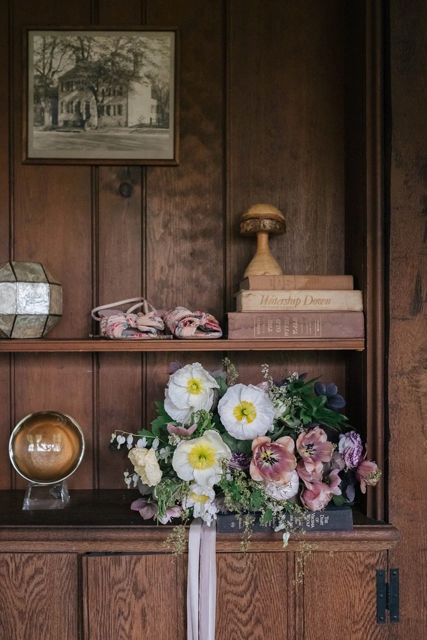 A wooden shelf with a vintage-style framed black and white photograph of a house with trees, a paper lantern, neatly folded cloth, a small mushroom-shaped wooden lamp, a stack of books, a round metallic decorative object, and a bouquet of white, pink, and purple flowers with greenery and ribbons.