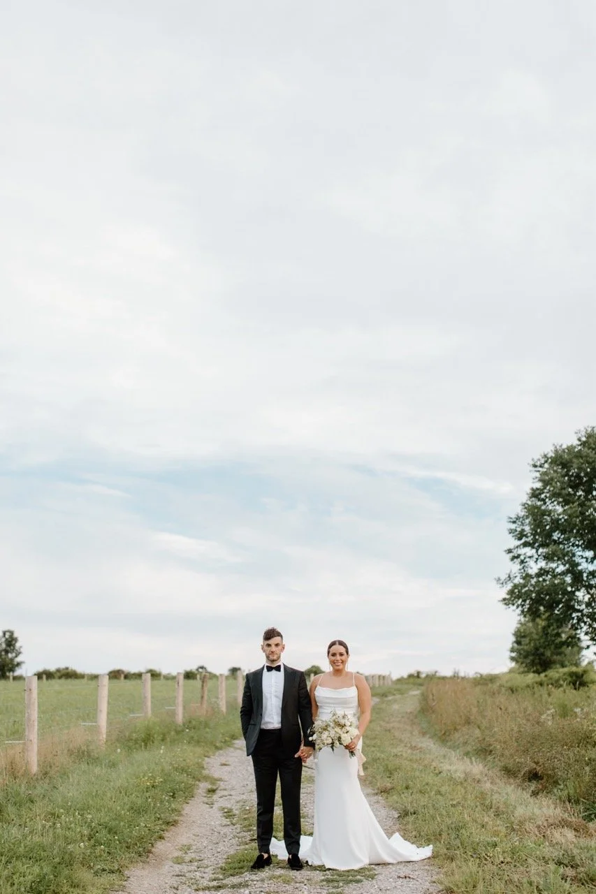 A bride and groom standing hand in hand on a dirt path in an open field, with a cloudy sky and trees in the background, dressed in wedding attire.