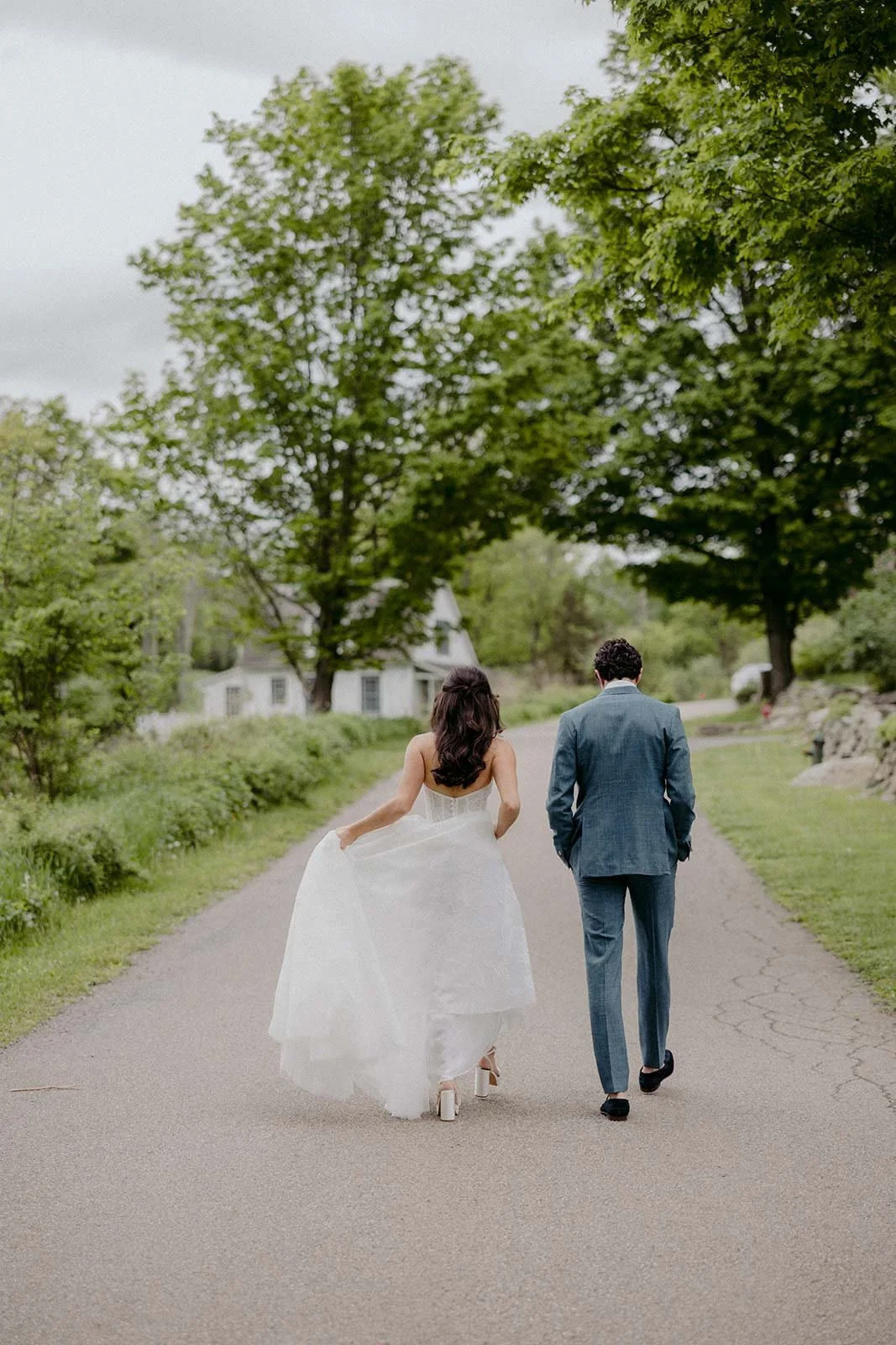 A bride and groom walking down a country road, surrounded by green trees and a white house in the background.