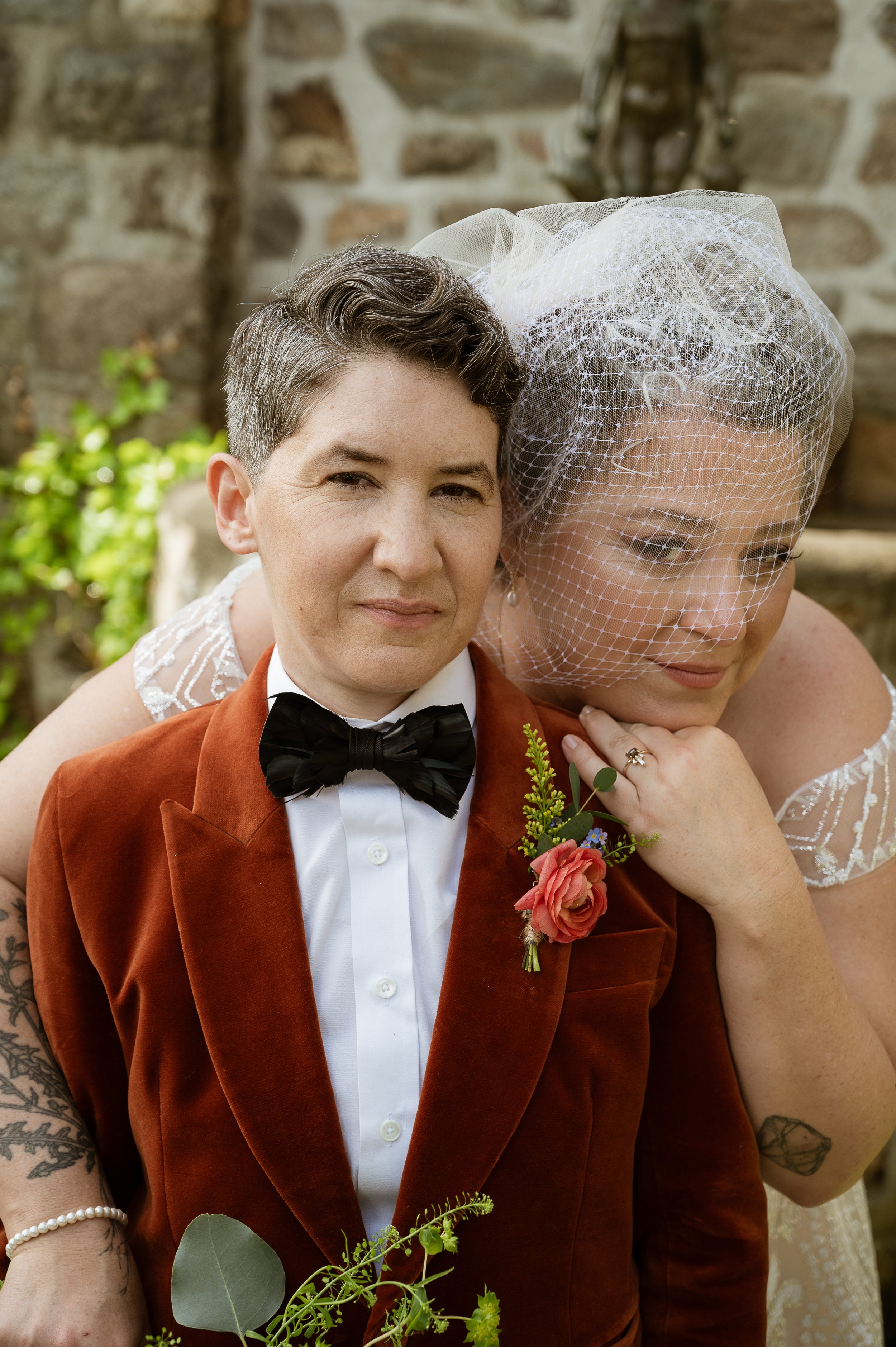 A person with short hair dressed in a tuxedo and bow tie, with a corsage, standing next to another person wearing a bridal veil, in front of a stone wall.