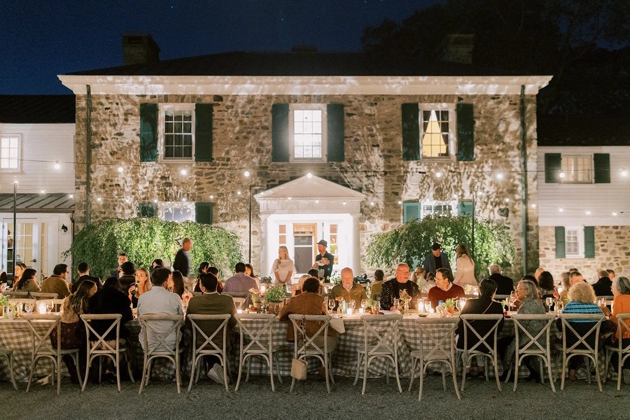 People dining at a long outdoor table with candles, in front of a stone house decorated with string lights at night.