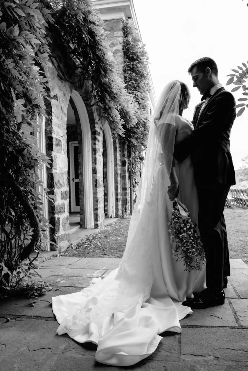 A bride and groom standing close together outdoors on their wedding day, with the bride's long train and veil visible, holding a bouquet of flowers, and smiling at each other.