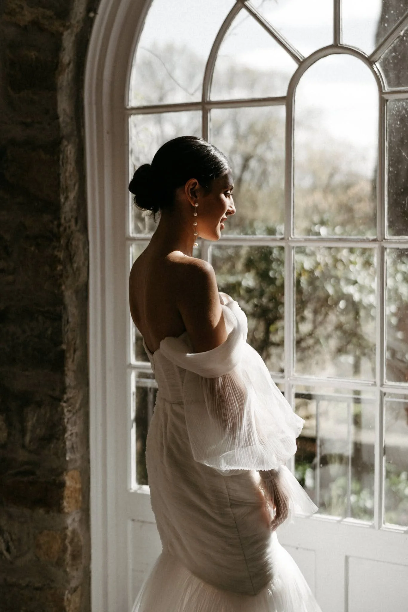 A woman in a white wedding dress with pearl earrings stands by a large window, looking outside with a serene expression.