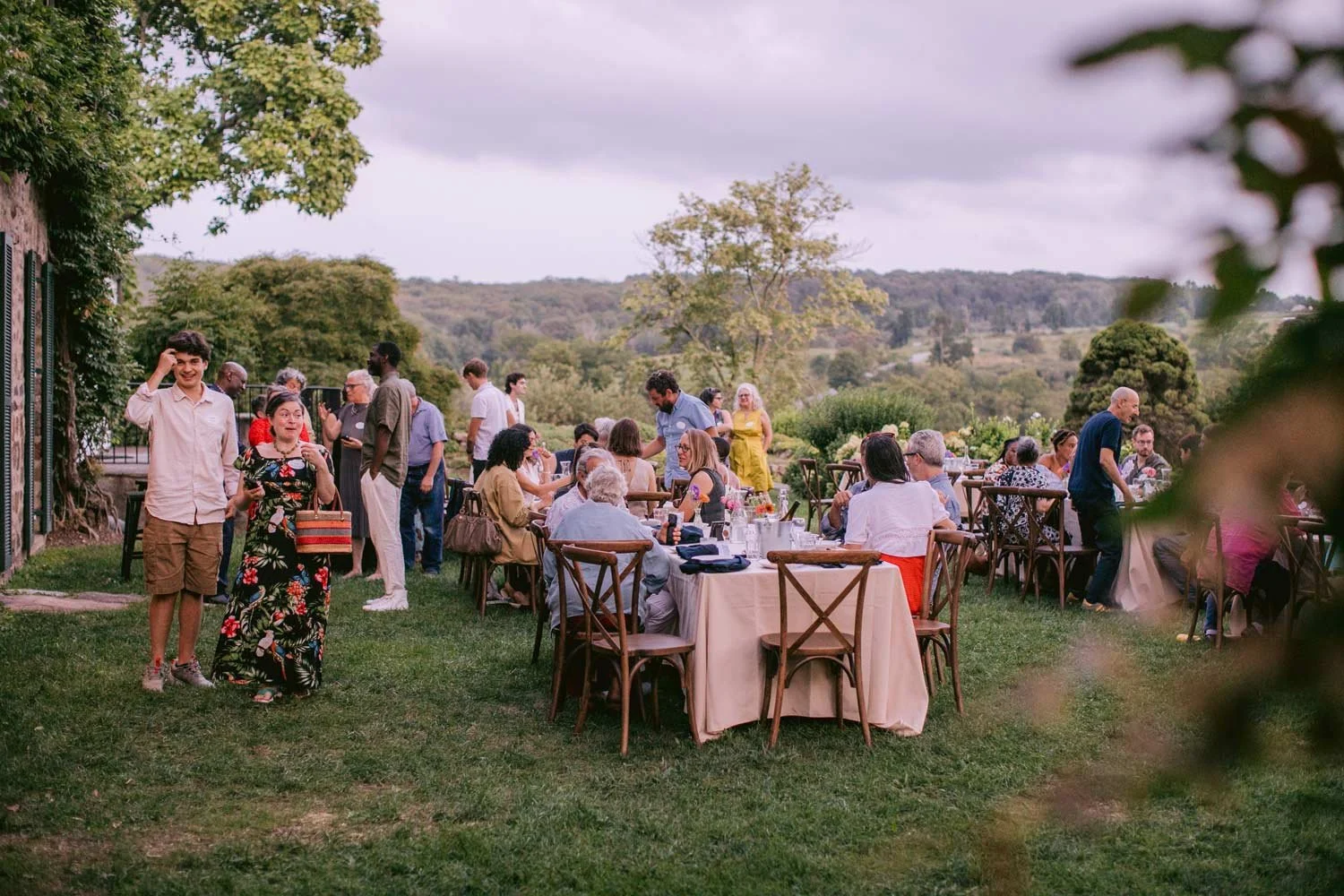 People dining and socializing at an outdoor gathering with tables and chairs on green grass, surrounded by trees and rolling hills in the background, during overcast weather.